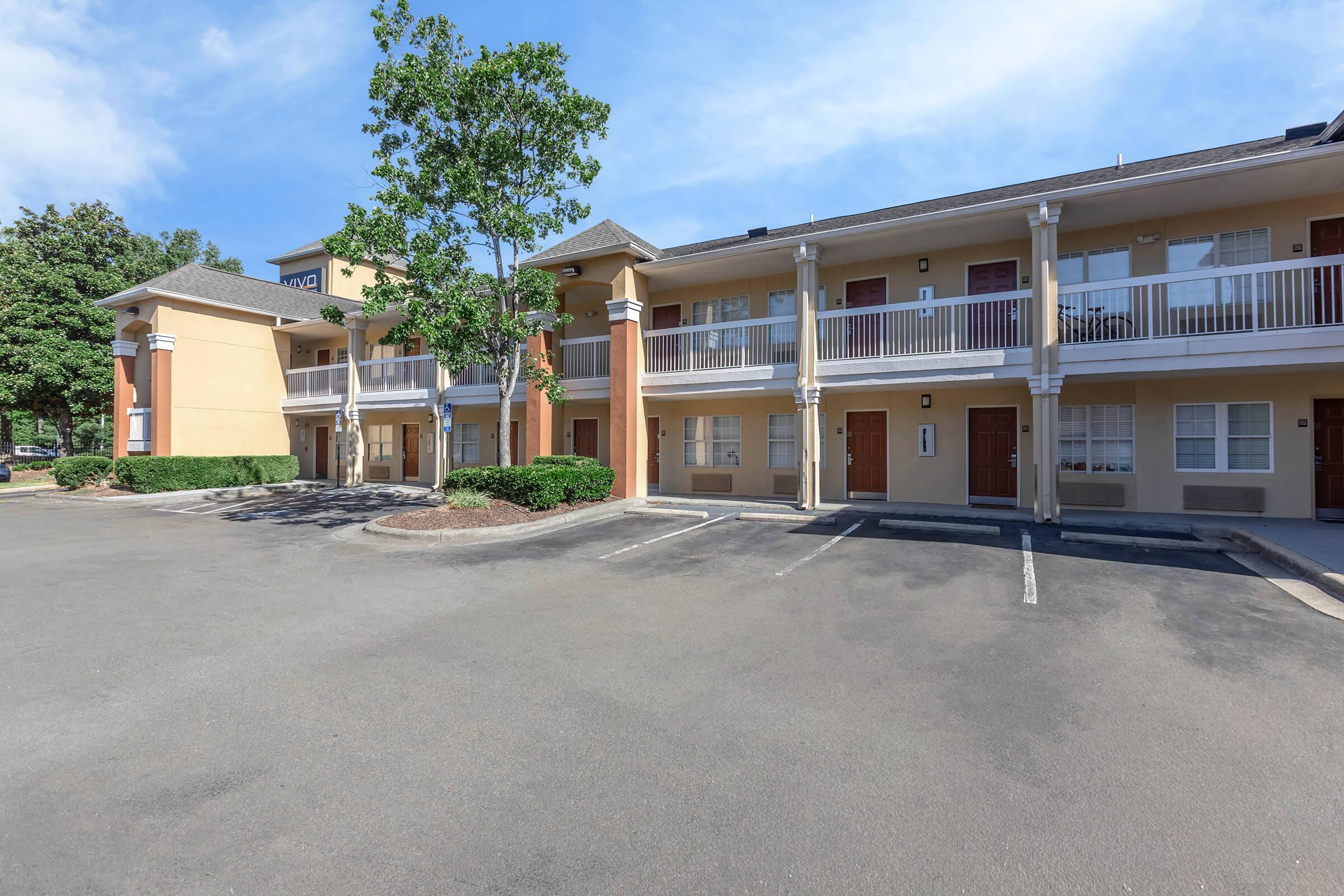 A view of a two-story hotel or apartment building with balconies on the upper level. The structure features beige and brown siding, surrounded by greenery and a parking lot. The sky is clear and blue, indicating a sunny day.