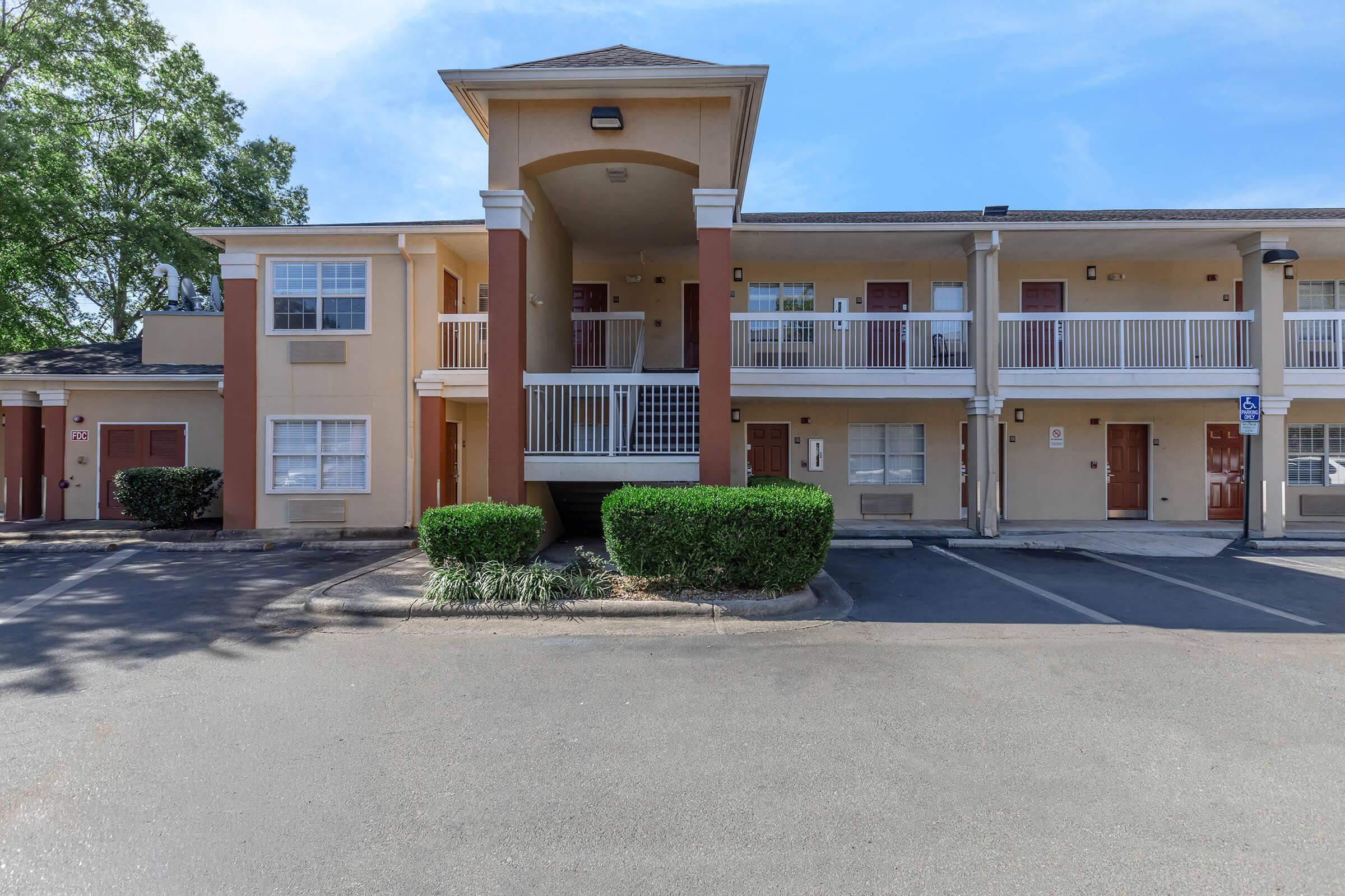 A two-story motel facade with a covered entrance. The building features a balcony on the upper level and several room entrances on the ground level, surrounded by landscaped greenery and a parking area. Clear blue sky visible overhead.