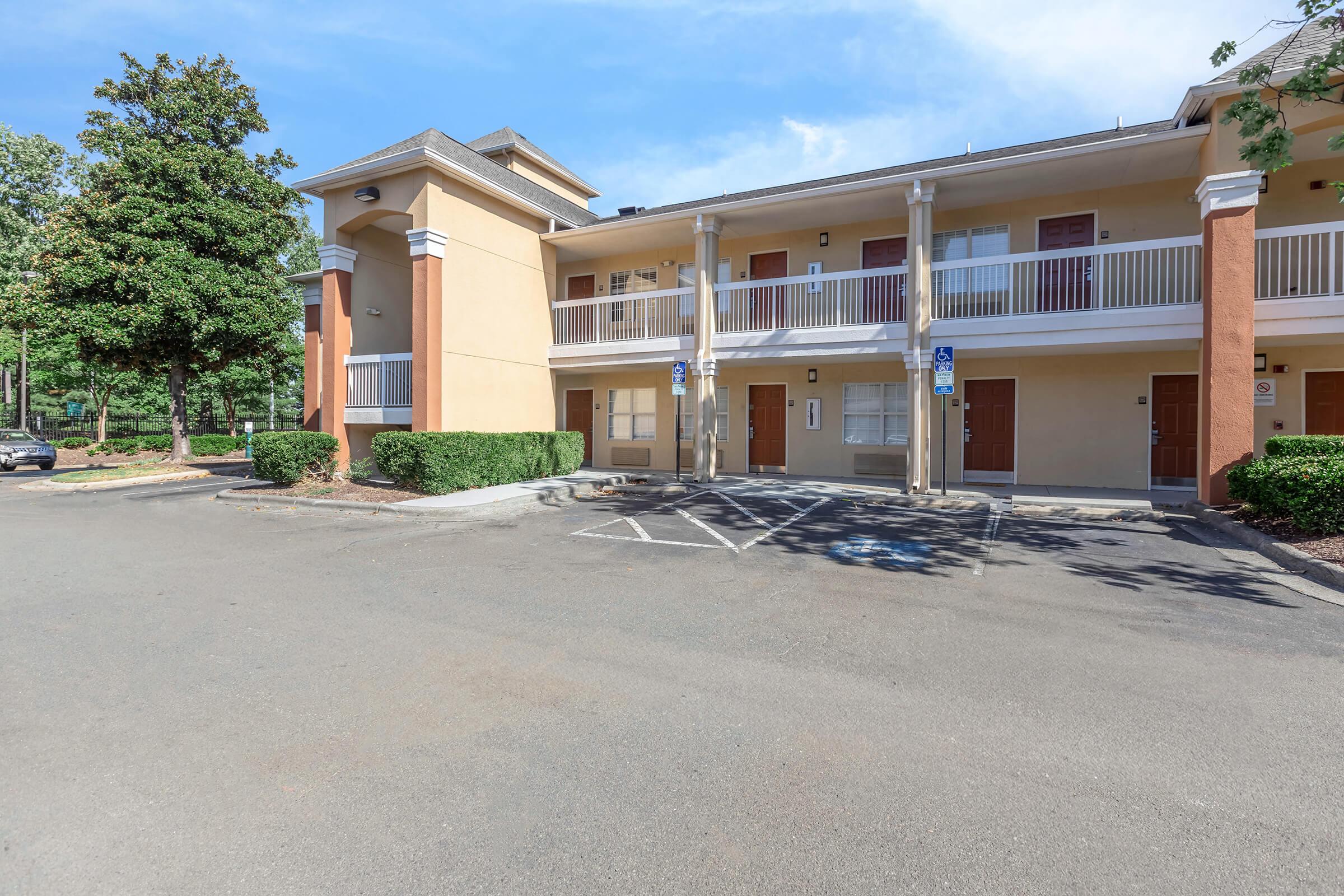 A view of a two-story hotel building with orange and beige exterior. It features white balconies, several parking spaces, and accessible parking spots in the foreground. Lush green trees are visible nearby, and the sky is clear with a few clouds.