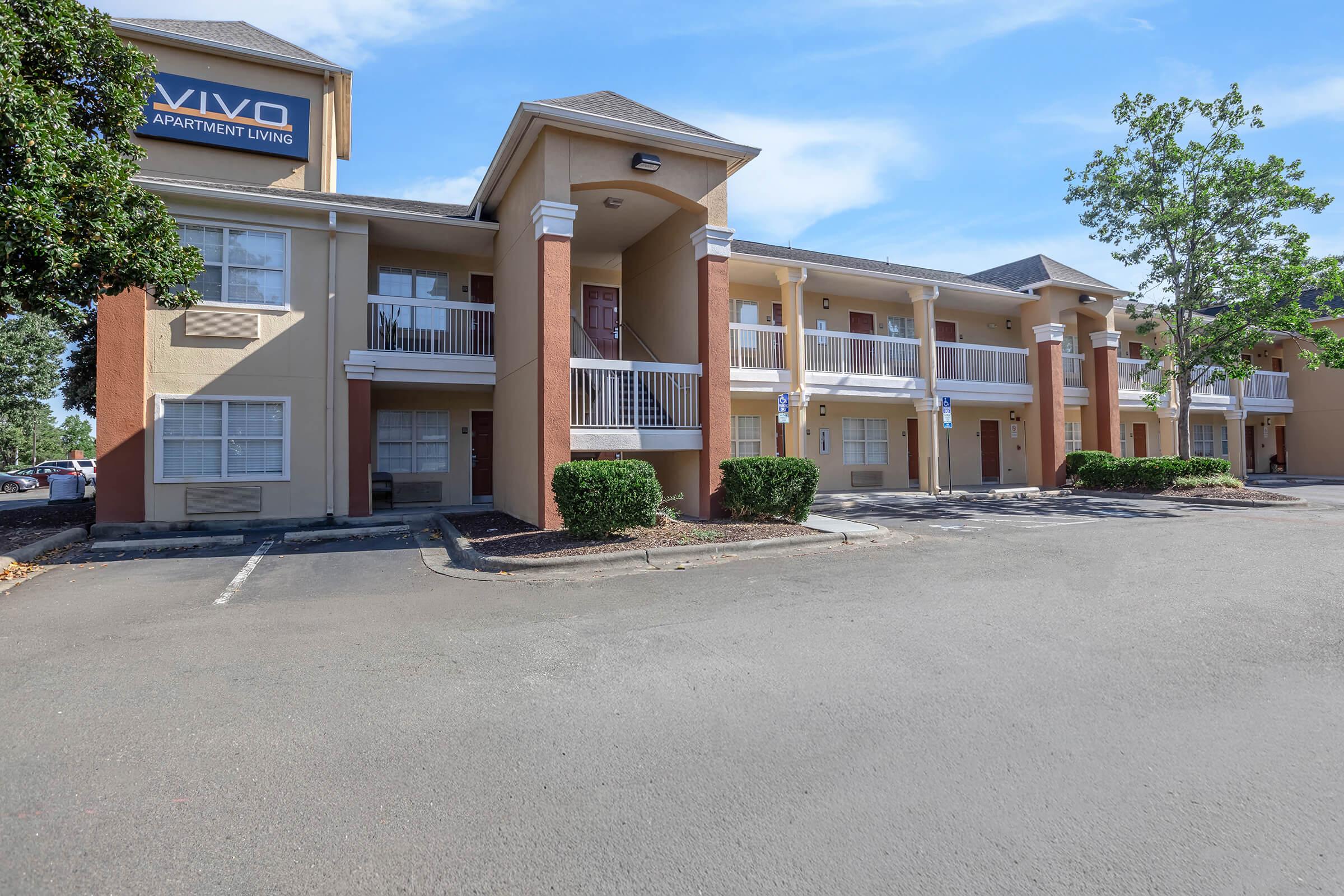 Exterior view of an apartment complex with a beige and orange color scheme. The building features multiple balconies, parking spaces, and landscaping with shrubs. A sign on the building indicates it is "Vivo Apartment Living." The sky is clear with some clouds.