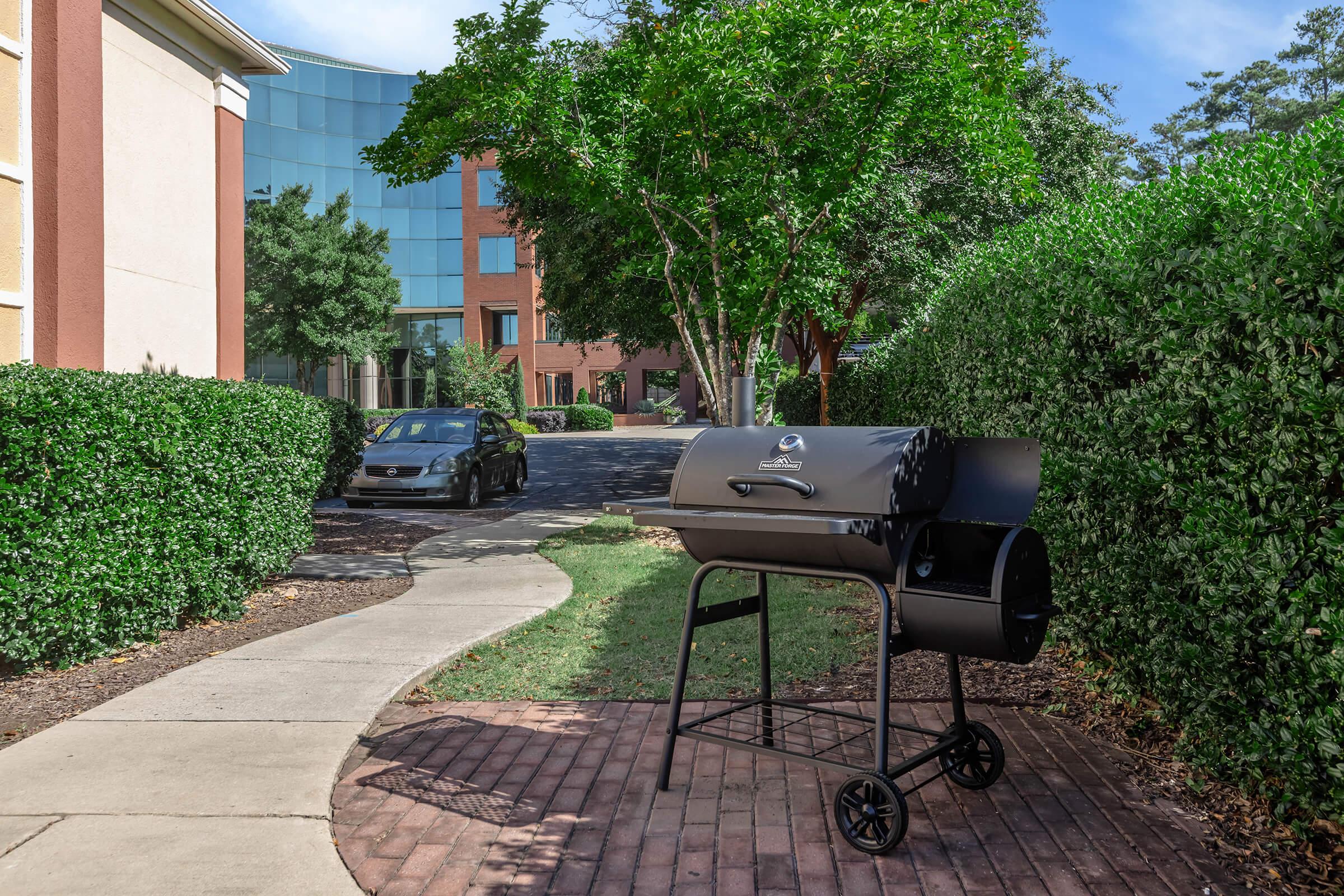 A black barbecue grill on a wheeled stand is positioned on a brick pathway surrounded by hedges. In the background, there is a tall glass building and a parked car nearby, with clear blue skies and trees providing some shade.