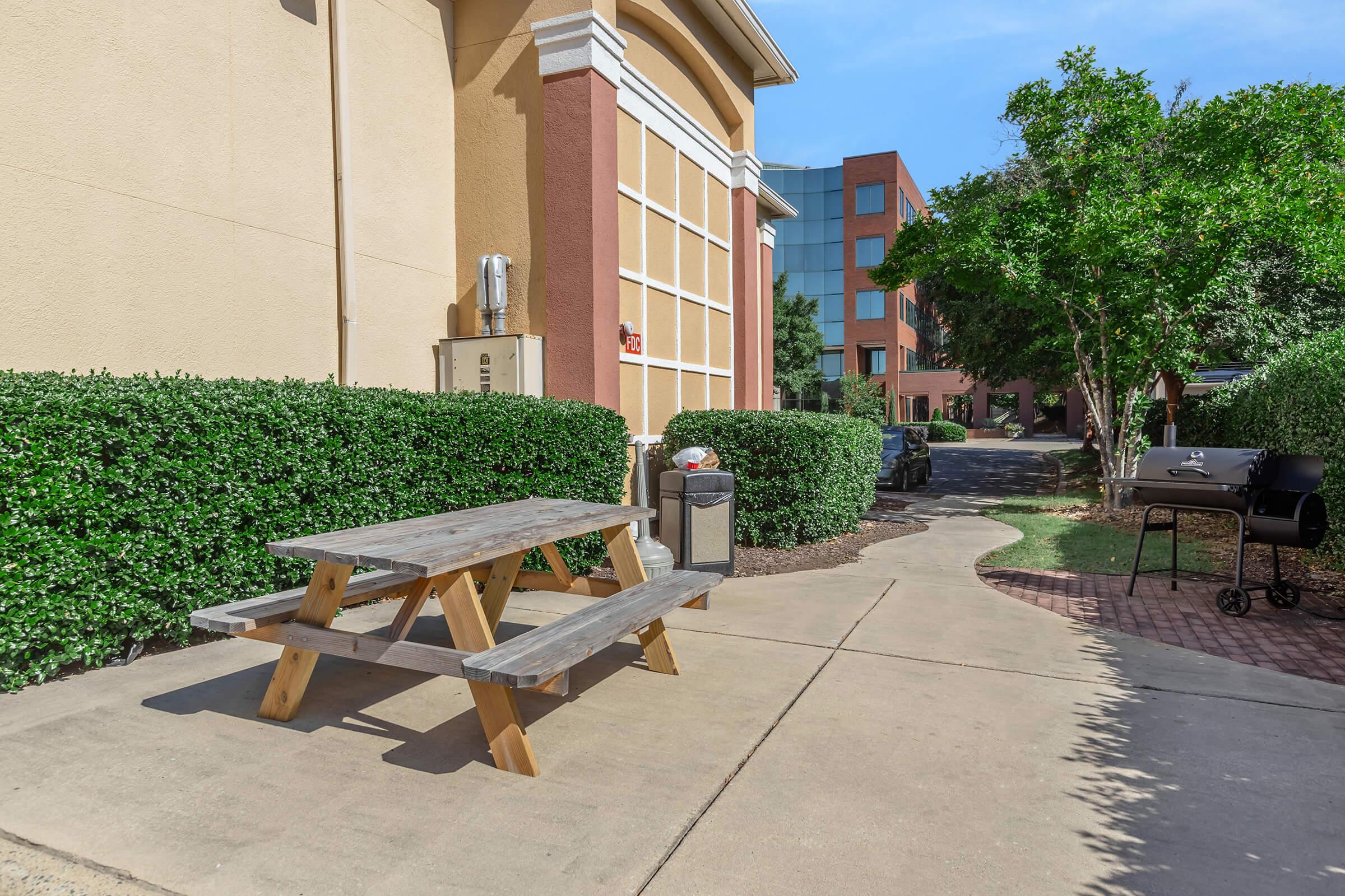 A small picnic area featuring a wooden table and benches on a concrete path. Nearby, there's a garbage can and a barbecue grill. Lush green bushes line the area, with a building and another paved path visible in the background under a clear blue sky.
