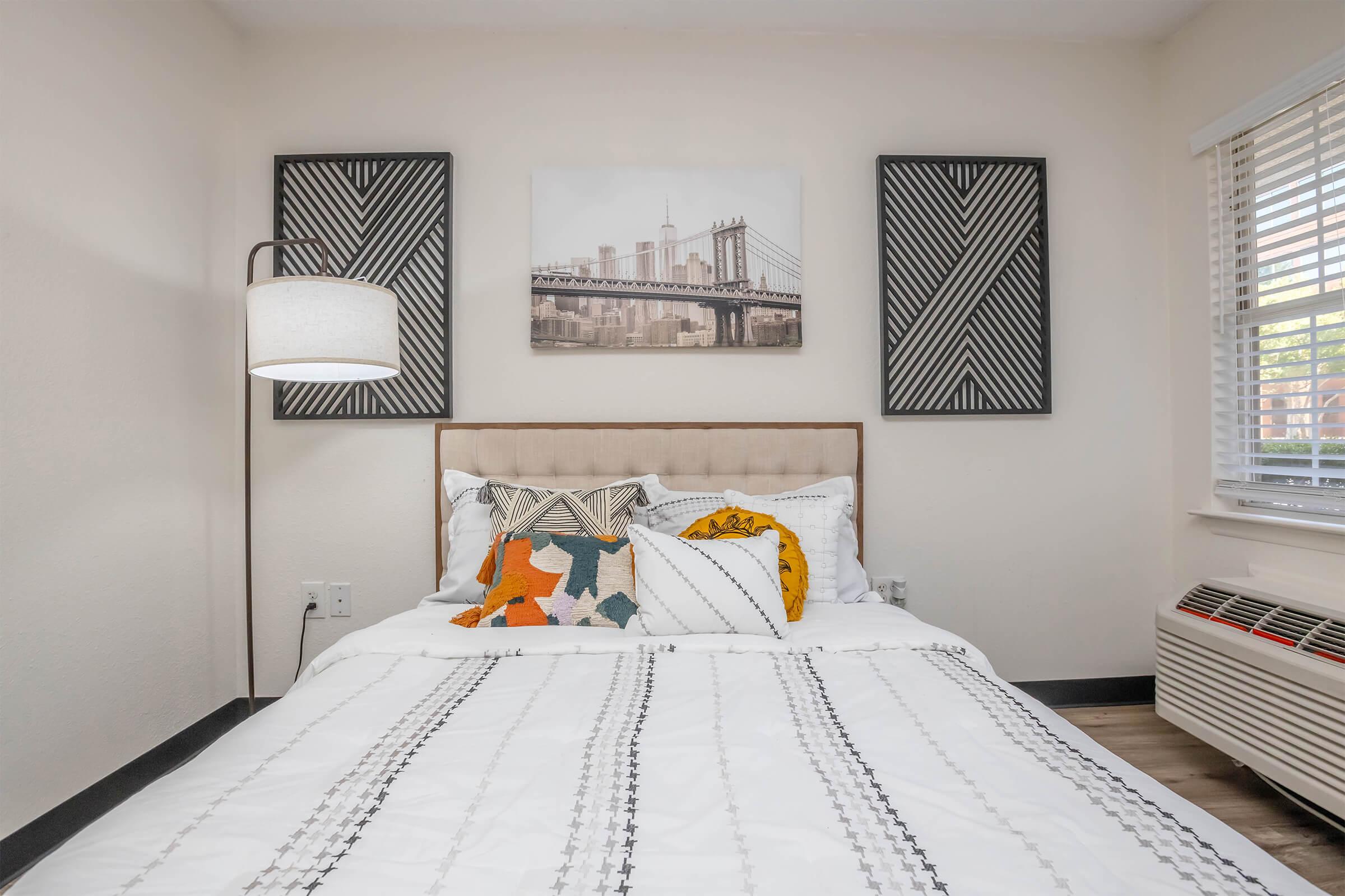 A cozy bedroom featuring a neatly made bed with decorative pillows, a modern lamp by the side, and artwork on the walls showcasing a city skyline. Natural light comes through the window, illuminating the warm, neutral tones of the room.