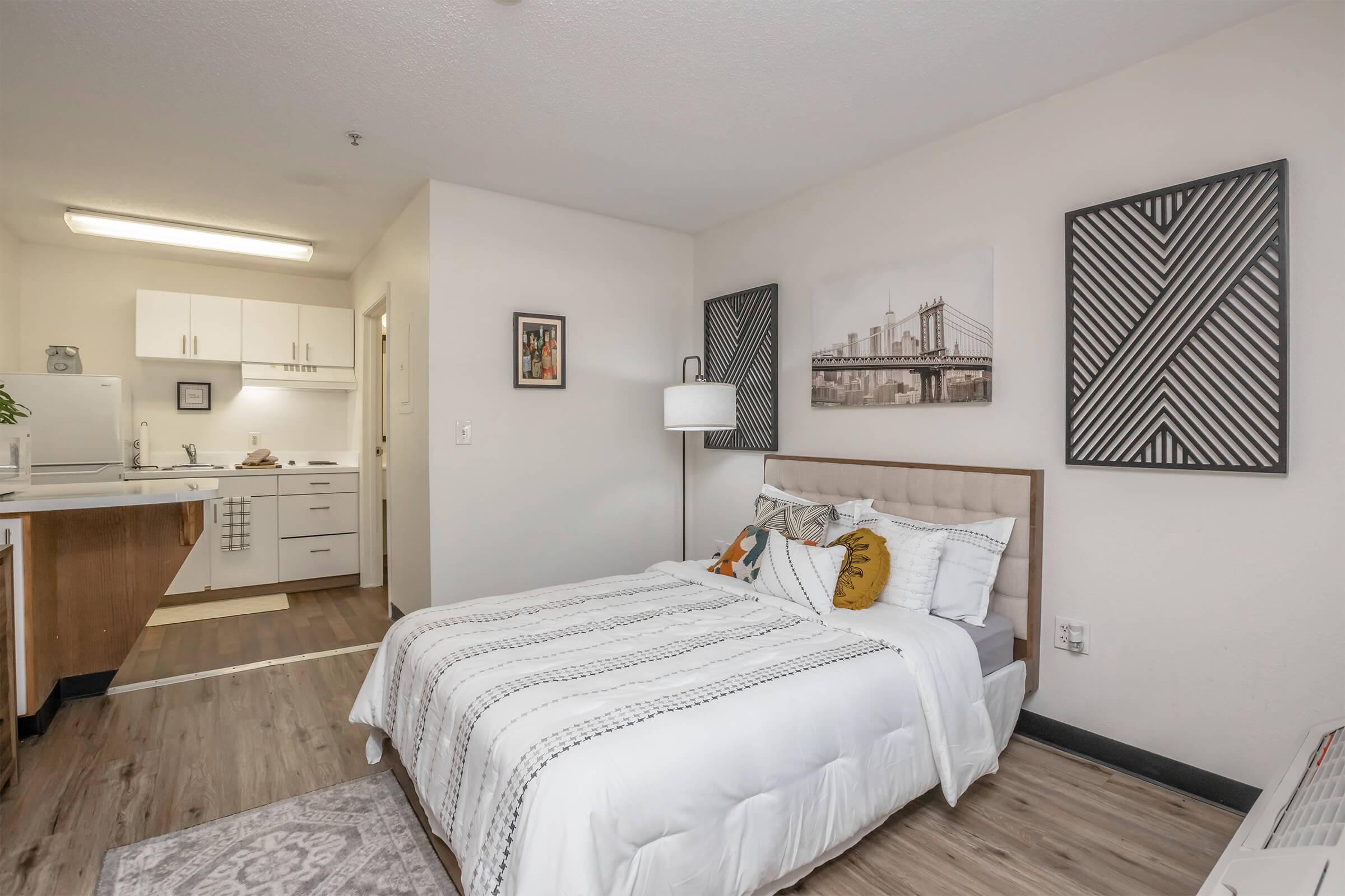 A cozy studio apartment featuring a neatly made bed with decorative pillows, a lamp beside it, and a small kitchen visible in the background. The walls are adorned with modern artwork, and there is a rug on the floor. The overall design is inviting and contemporary.