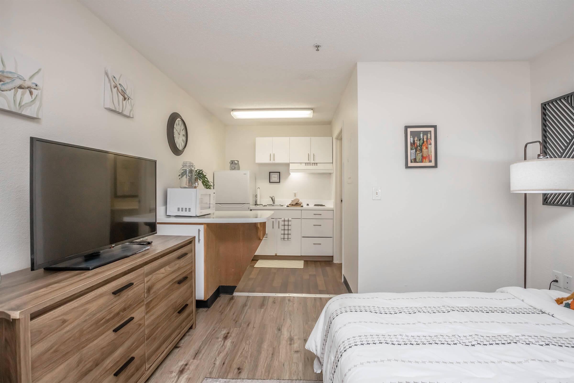 A cozy studio apartment featuring a kitchen area with white cabinets and a dining space. A wooden TV stand holds a large television, while a bed with a striped blanket sits nearby. The walls are adorned with simple artwork, and a lamp stands next to the bed, creating a warm and inviting atmosphere.