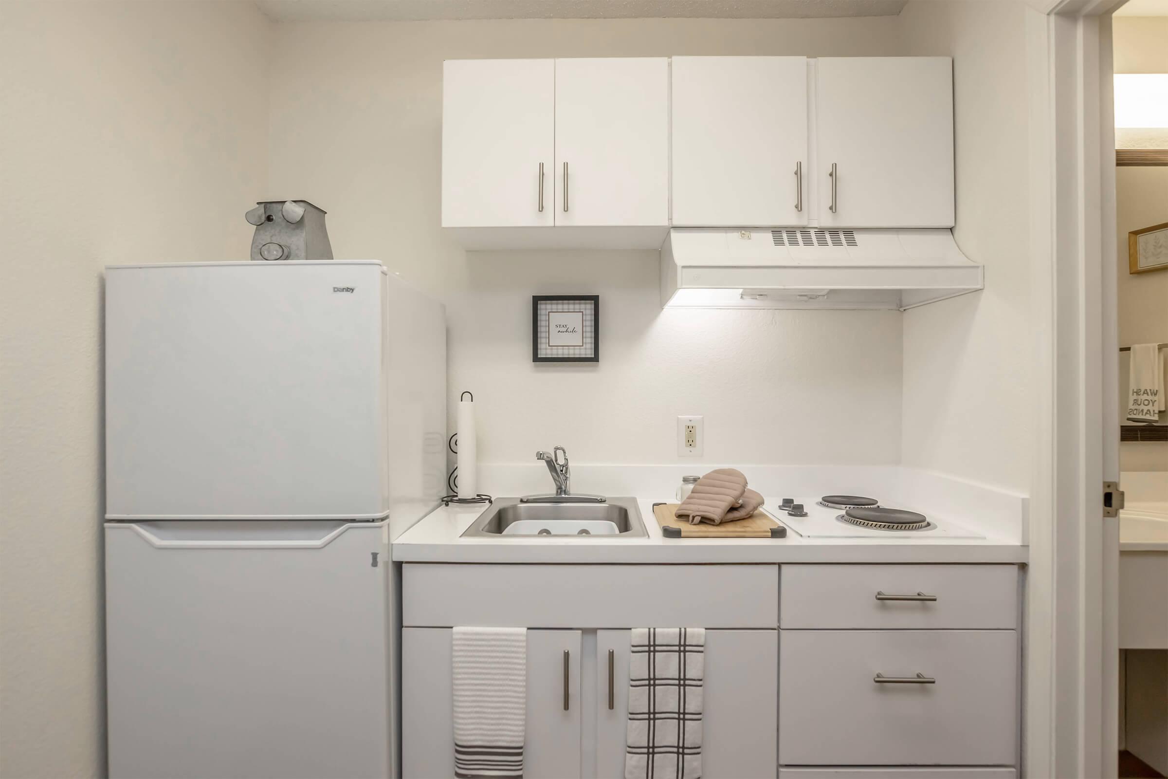 A modern kitchen featuring a white refrigerator, sink, and stove. The cabinetry is sleek and minimalist, with a towel hanging from the handle. A coffee maker is placed on the countertop, along with a cutting board. The overall design is clean and bright, enhancing the spacious feel of the area.