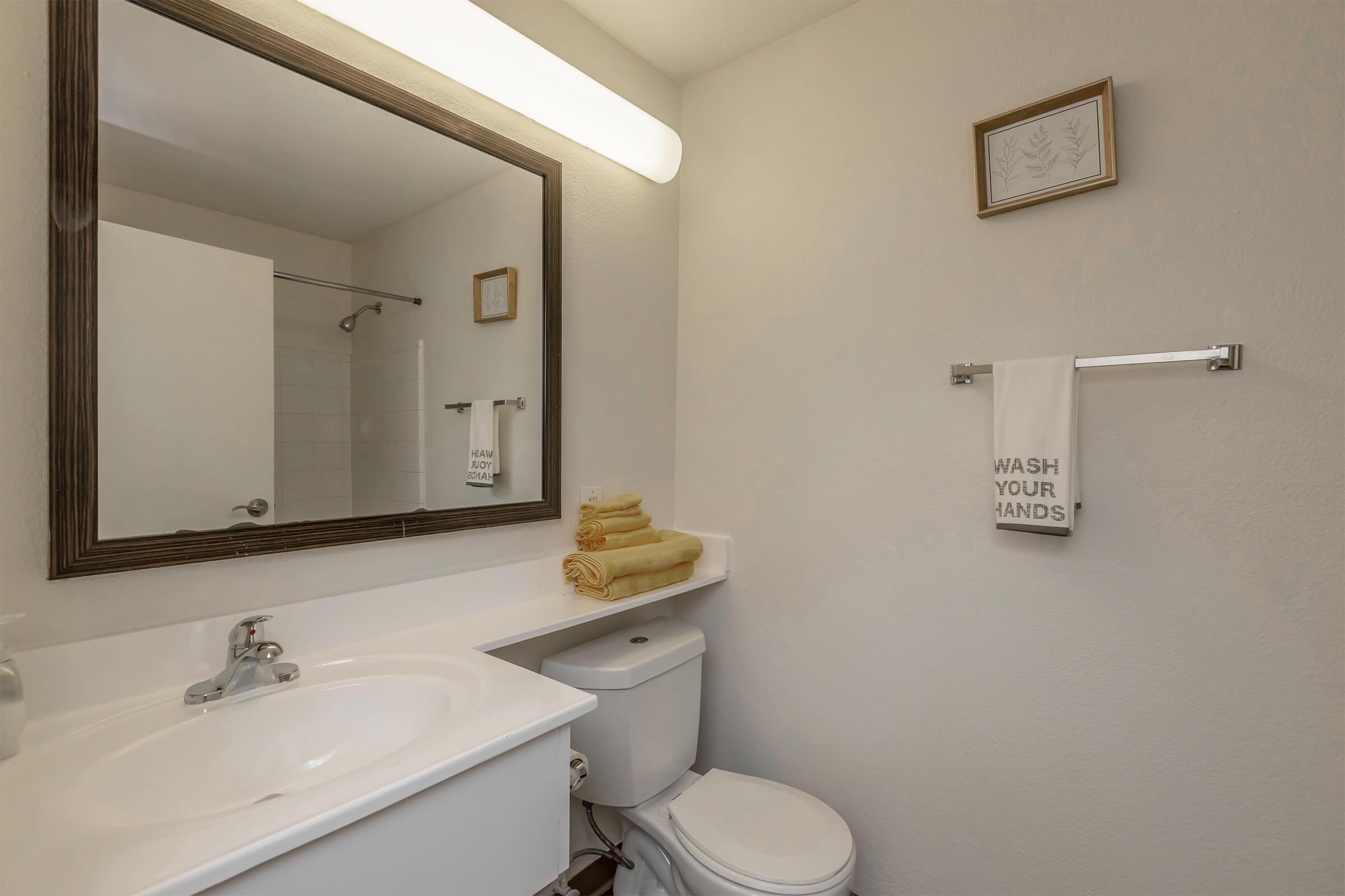 A clean bathroom with a large mirror above a sink, a towel rack holding a towel with the text "WASH YOUR HANDS," and neatly stacked yellow towels. A small framed item is on the wall, and a shower area is visible in the background. The overall decor is simple and modern.