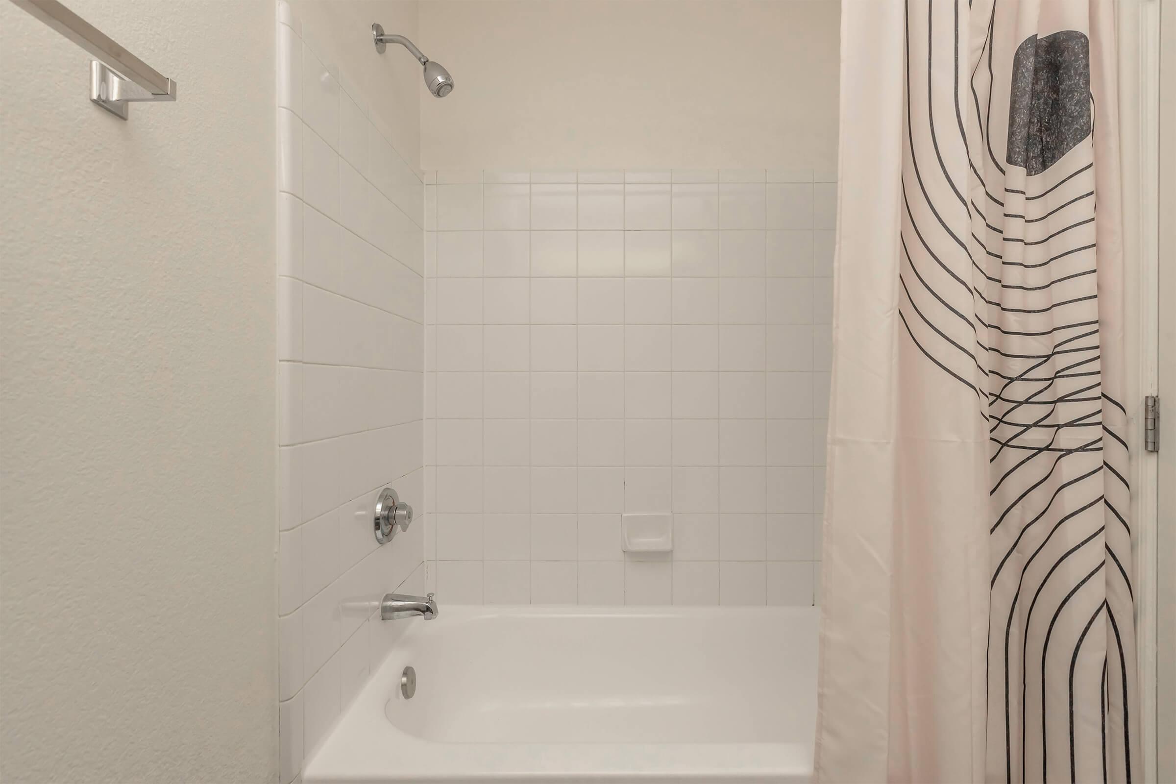 A clean bathroom featuring a white bathtub with a modern showerhead, surrounded by white tiled walls. A light-colored shower curtain with black lines hangs in front of the tub, adding a subtle design element. The space is simple and bright, conveying a fresh and minimalist aesthetic.