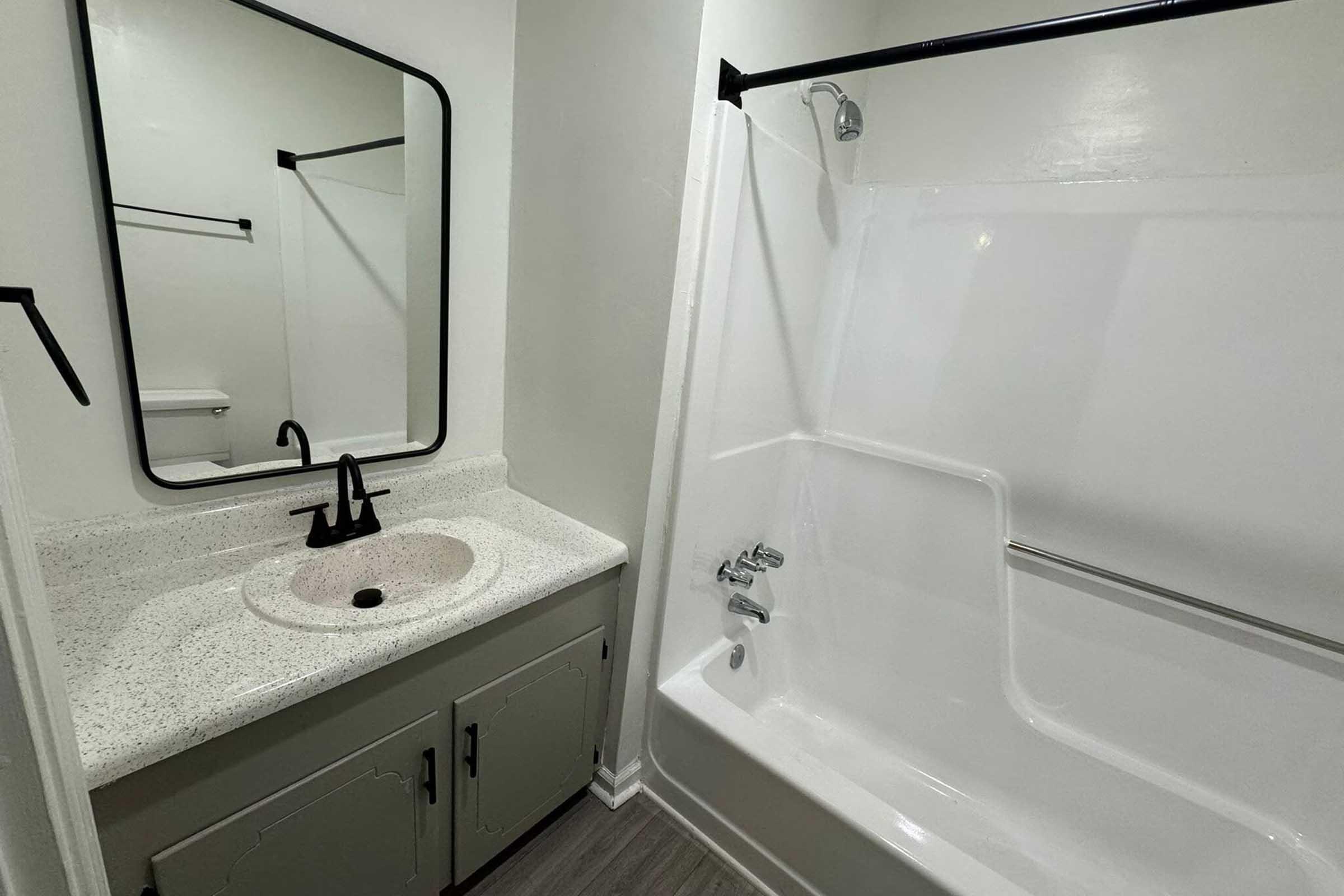 A clean bathroom featuring a white bathtub and shower combination, a light-colored vanity with a black faucet, and a round mirror with a black frame. The walls are painted white, and the flooring is a light gray wood-like material. A towel rack is visible on the left.