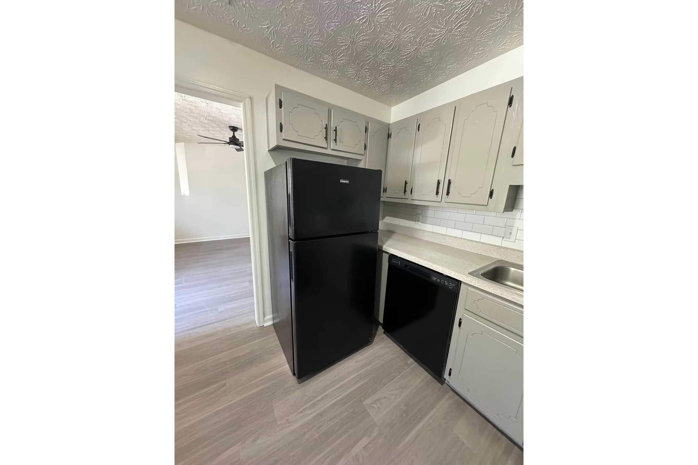 A kitchen featuring a black refrigerator and a black dishwasher next to light-colored cabinets and a countertop. The flooring is a light wood tone, and there's a doorway leading to another room in the background, with a ceiling fan visible.