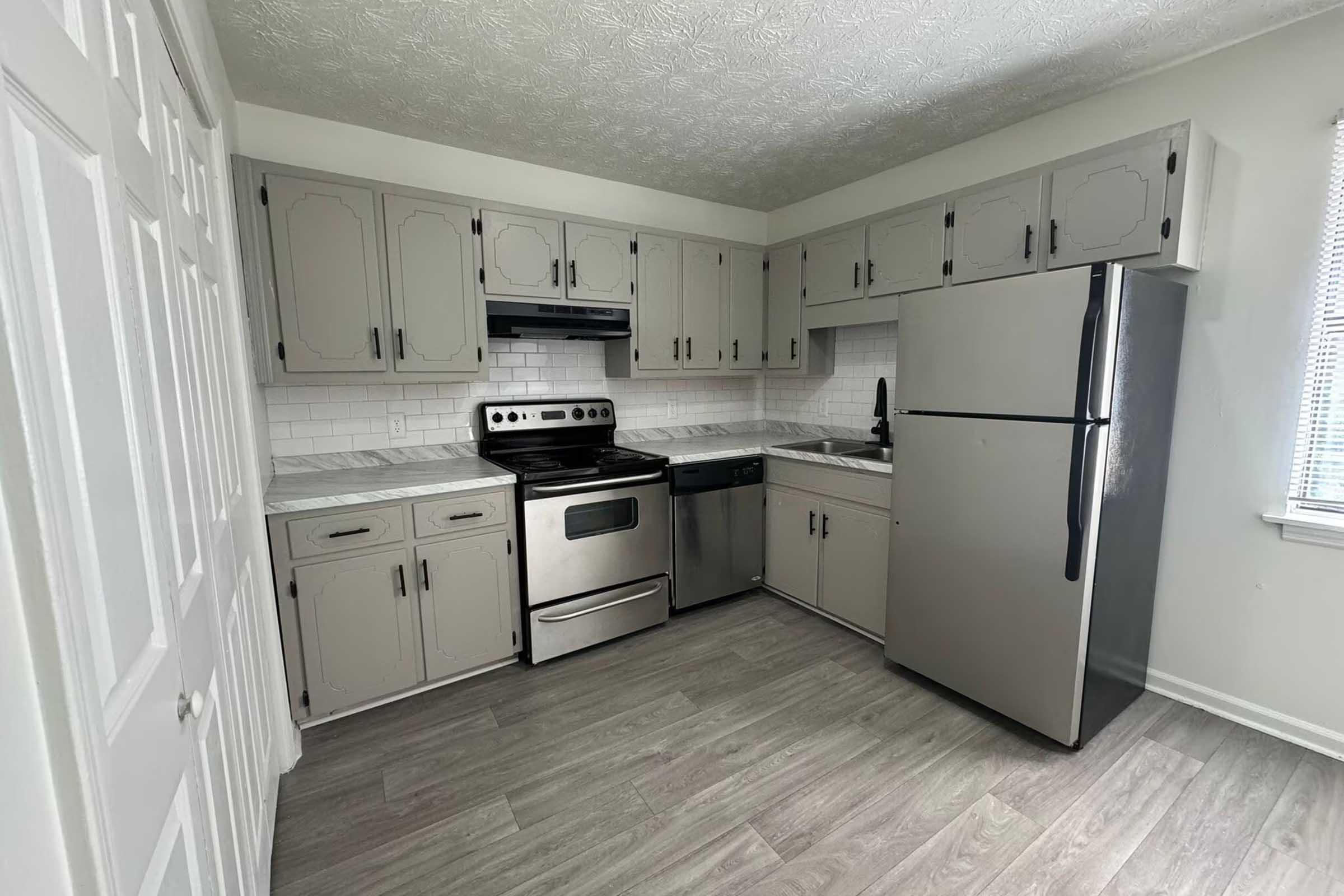 A modern kitchen featuring light gray cabinetry, stainless steel appliances including a refrigerator, oven, and dishwasher, and white tiled backsplash. The flooring is gray laminate, and there is a window providing natural light. Cabinets are above the counter and appliances.