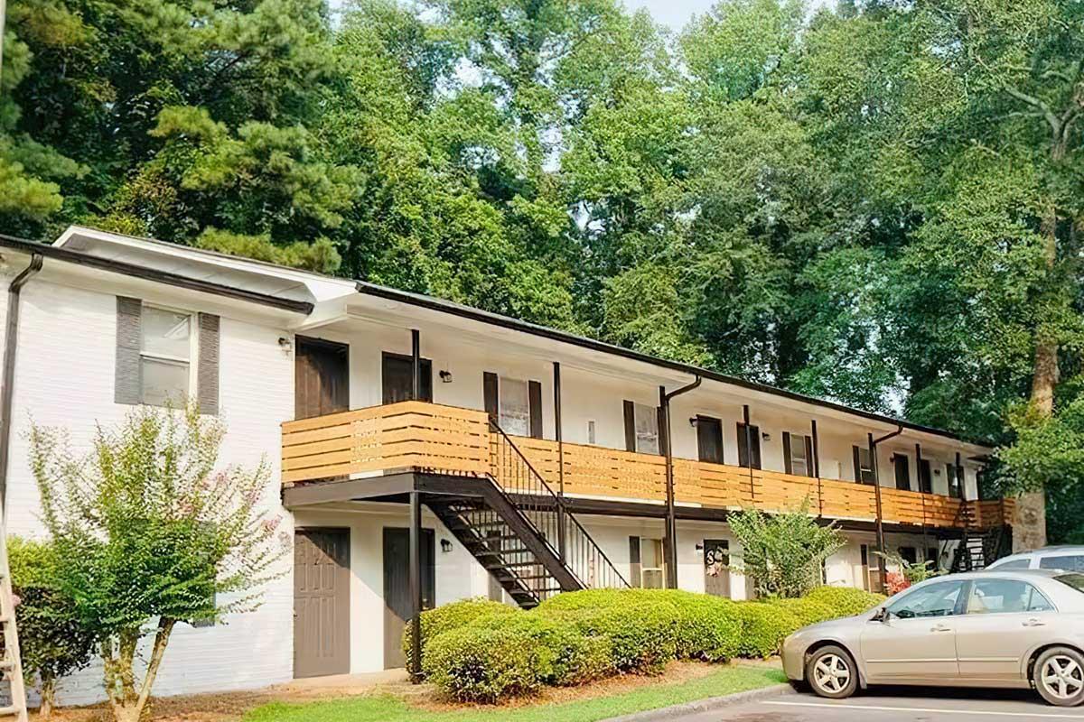 A two-story apartment building with wooden balconies, surrounded by greenery and trees. The building has a clean, white exterior with dark-colored doors and railings. A parking area is visible in front, with a car parked nearby and manicured shrubs lining the walkway.