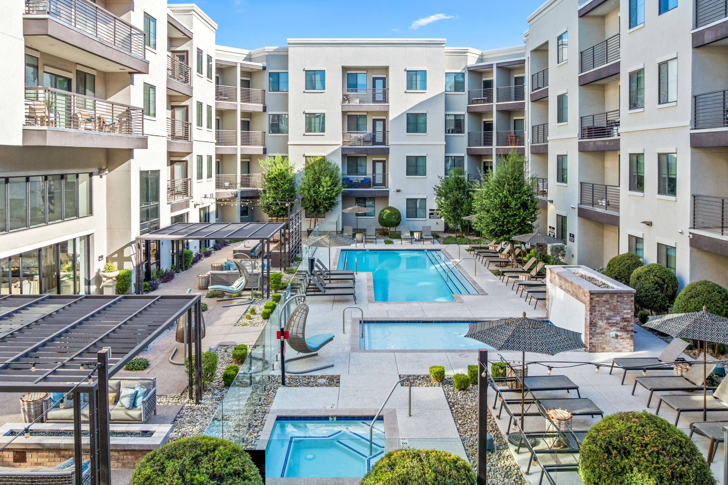 A view of a modern apartment complex courtyard featuring two swimming pools surrounded by lounge chairs, shaded cabanas, and neatly manicured bushes. The buildings have balconies, and there are seating areas with outdoor furniture, creating a resort-like atmosphere.