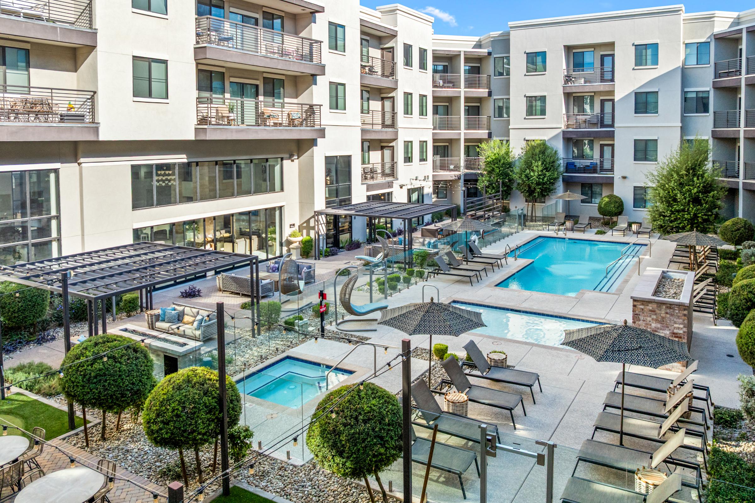 A spacious outdoor pool area surrounded by modern apartment buildings. Two swimming pools, lounge chairs, and shaded cabanas are visible. Lush greenery and decorative landscaping enhance the aesthetic. Some residents are enjoying the space, creating a relaxed and inviting atmosphere.