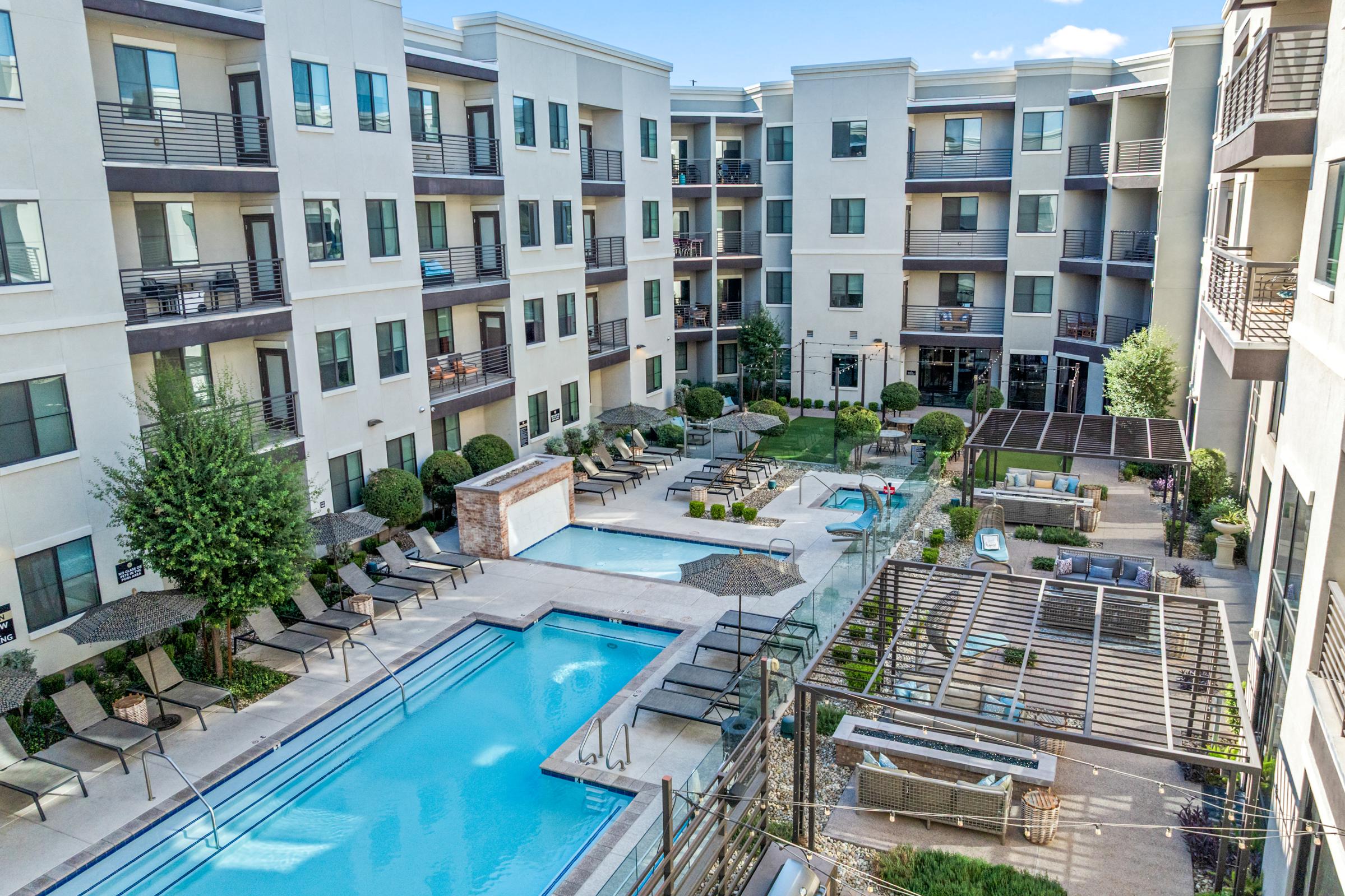 Aerial view of a modern apartment complex featuring a swimming pool, lounge areas with chairs, decorative landscaping, and balconies. The design includes a mix of natural and built spaces, with greenery around the pool area and outdoor seating for residents.