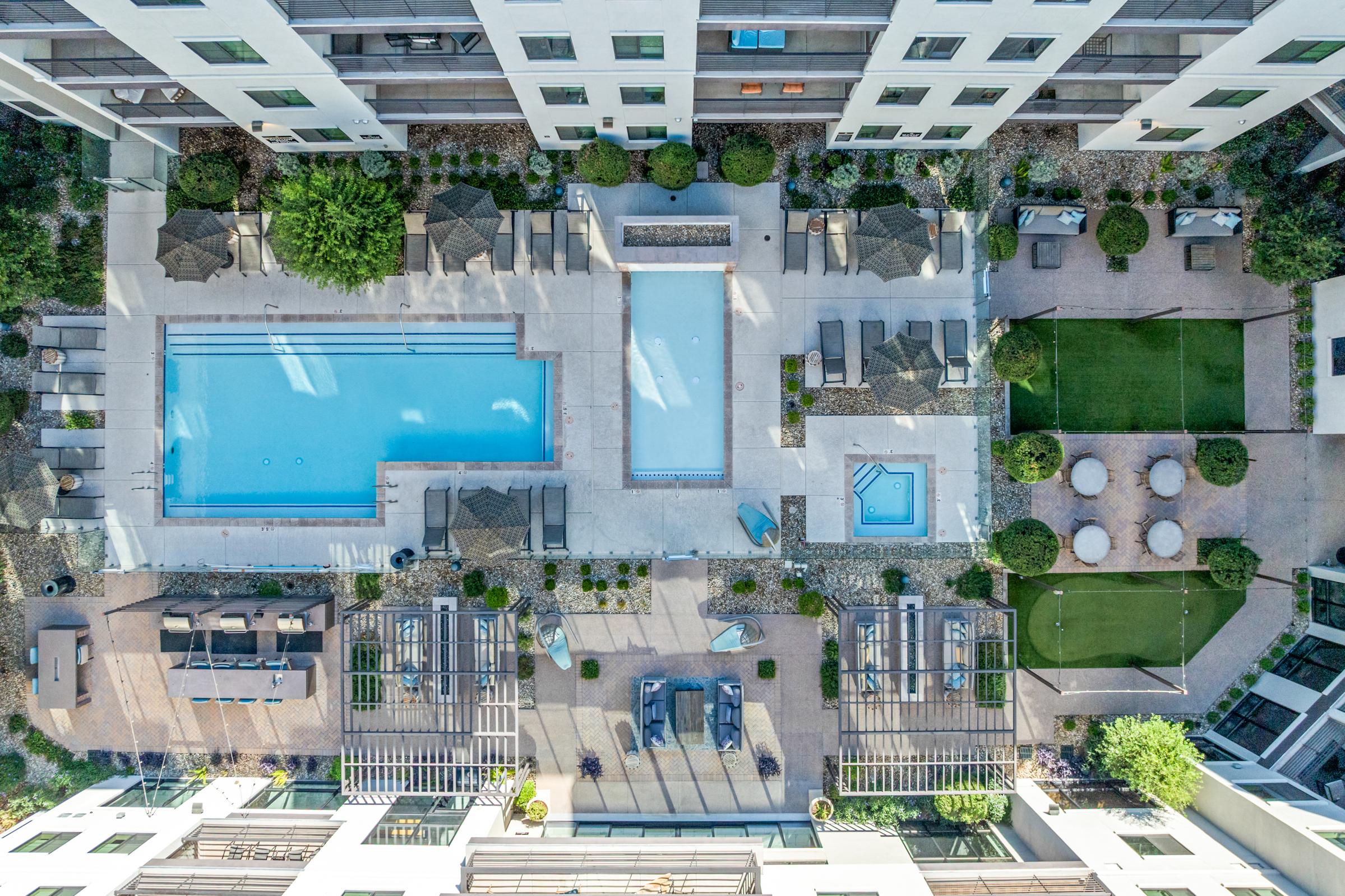 Aerial view of a modern apartment complex featuring a large swimming pool, a hot tub, and surrounding lounge areas with chairs and shaded spots. The pool area is landscaped with greenery and neatly arranged furniture, including sun loungers. Well-maintained lawns and seating arrangements are visible, creating an inviting outdoor space.