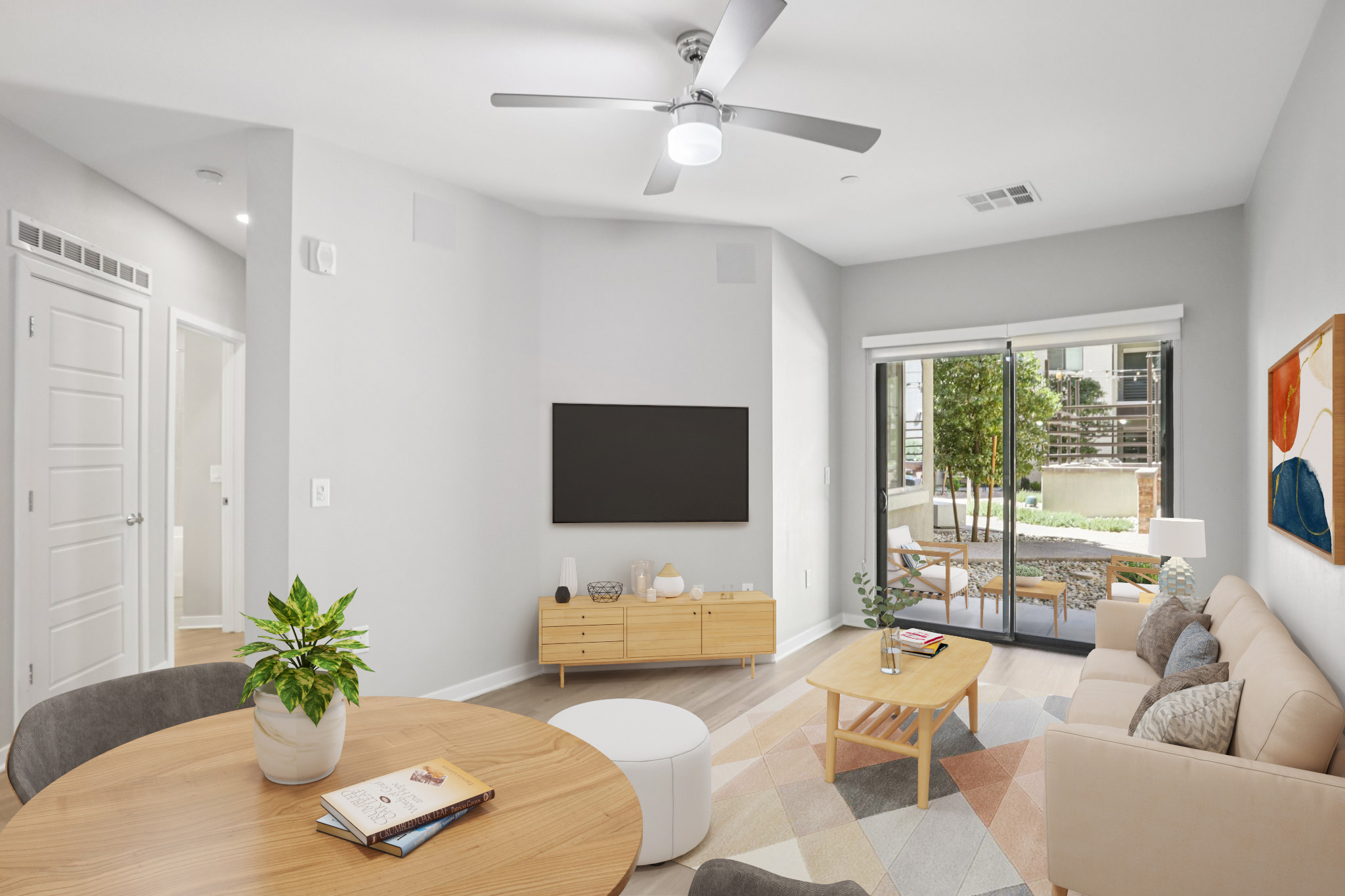 A modern living room featuring a beige sofa, a wooden coffee table, and a round dining table. There is a television mounted on the wall, a ceiling fan, and large windows with a view of a patio. Decorative items include a plant and art on the wall, creating a bright and inviting atmosphere.