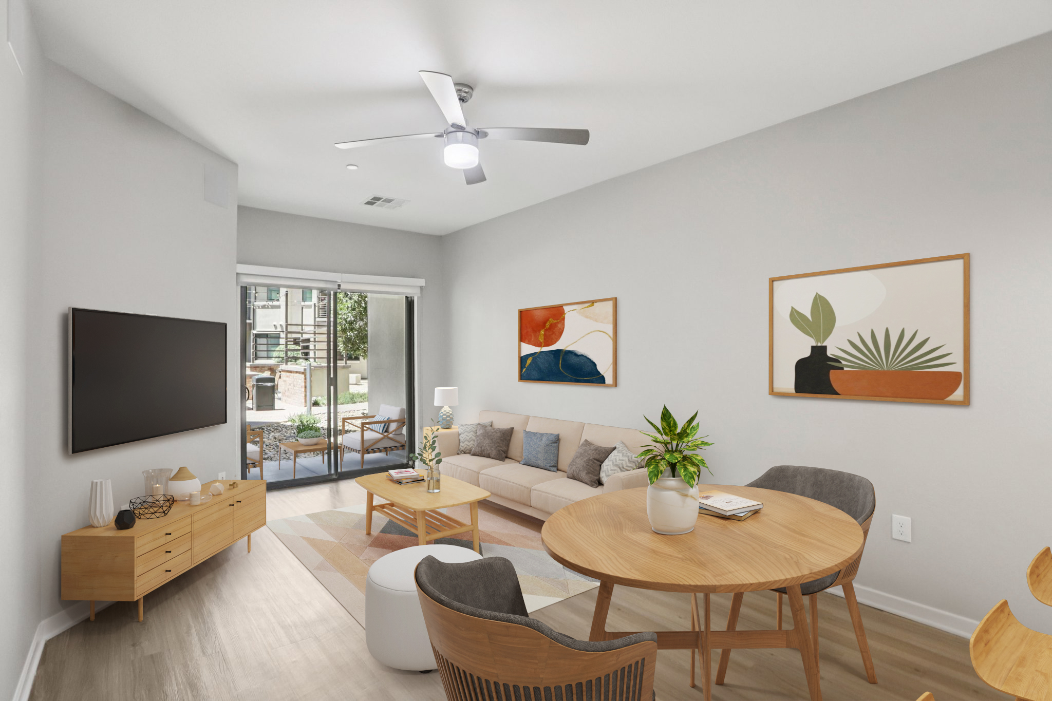 A modern living room featuring a light-colored sofa, wooden furniture, and a round dining table. There are decorative wall art pieces and a flat-screen TV. Large windows let in natural light, showcasing a view of an outdoor patio with seating. The decor includes plants and a neutral color palette.