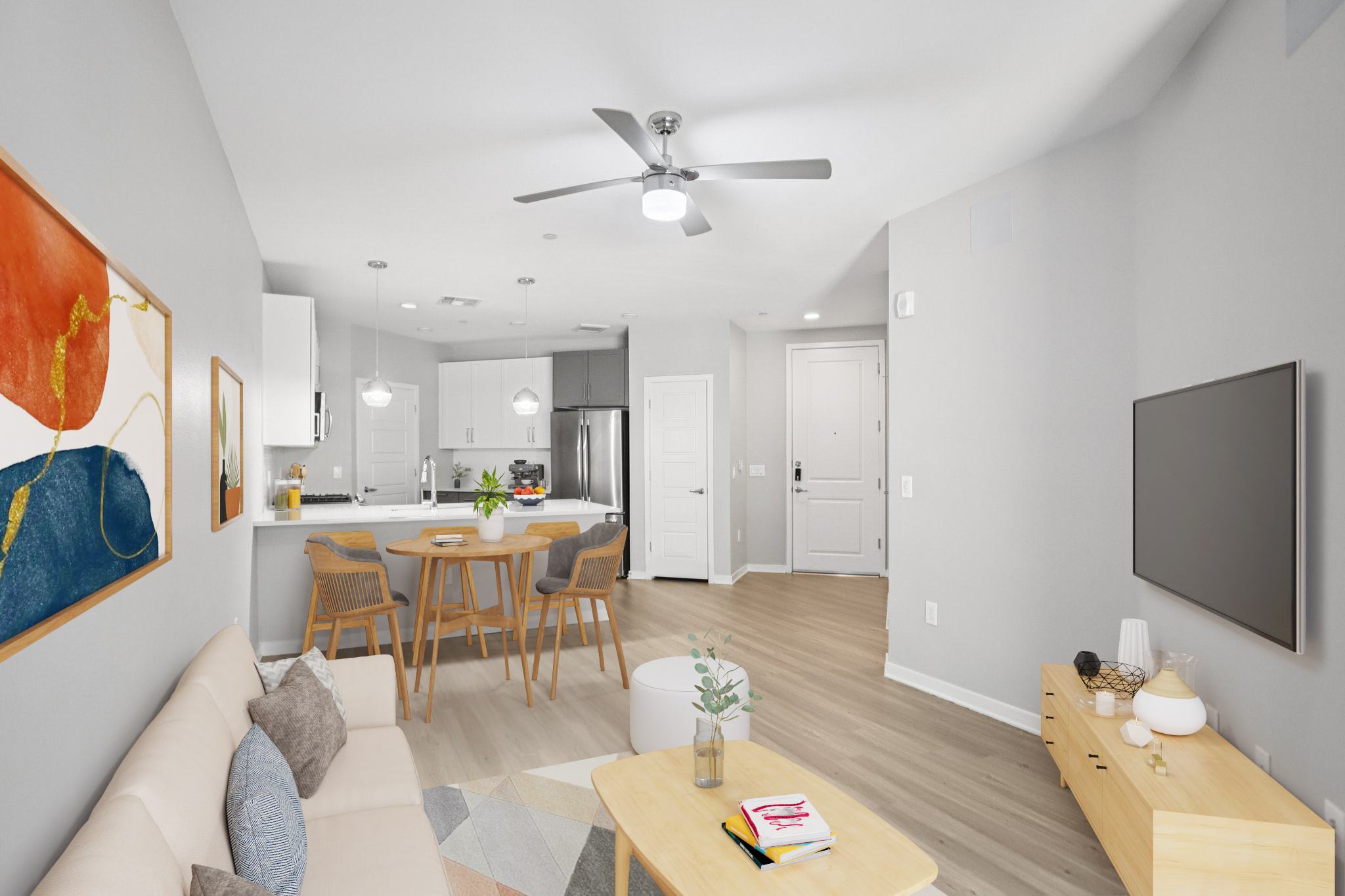 Modern living room and kitchen area in a stylish apartment. Features a light-colored sofa, wooden dining table with chairs, decorative art on the wall, and a ceiling fan. The space has neutral gray walls, light wood flooring, and a cozy, inviting atmosphere.