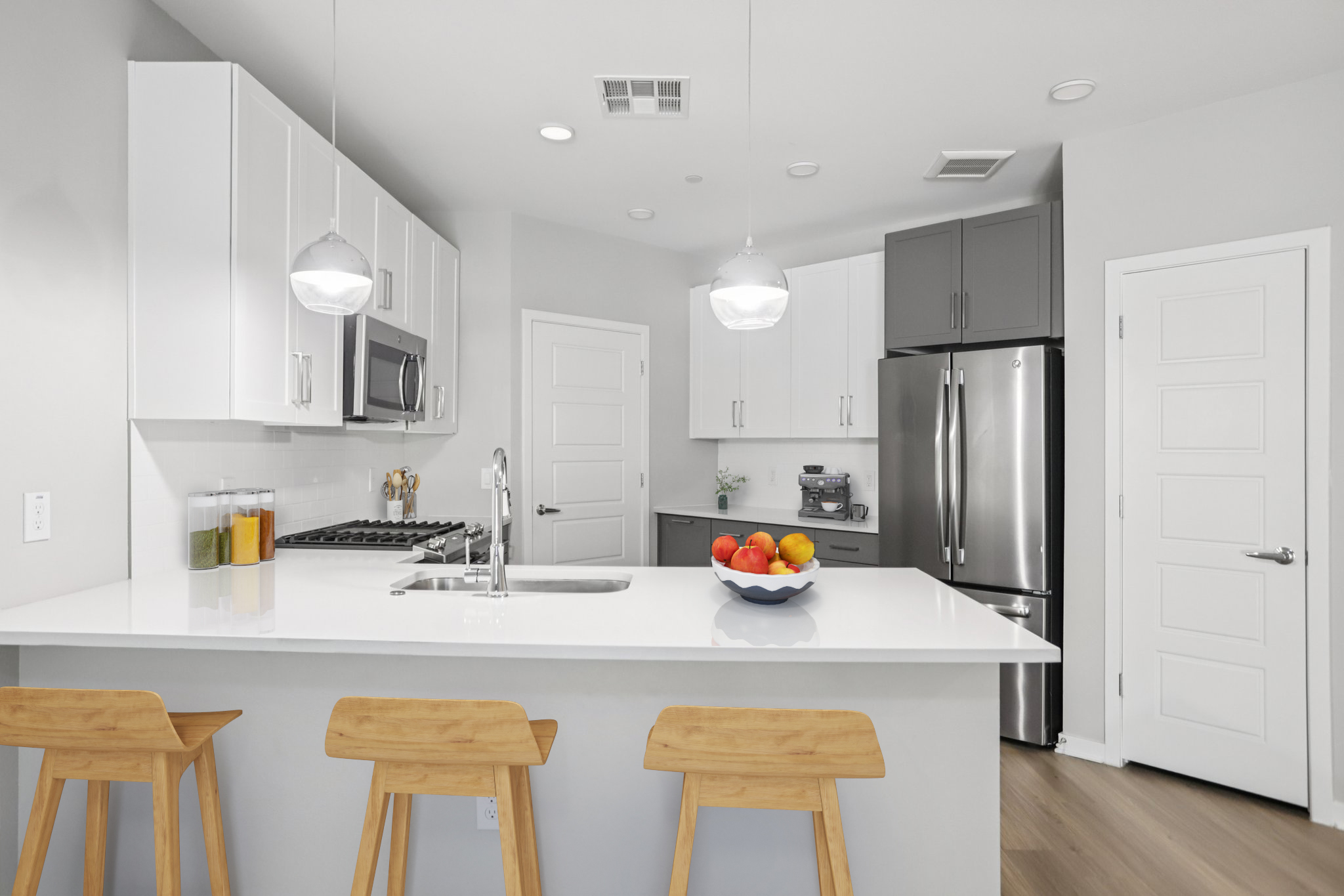 Modern kitchen featuring white and gray cabinetry, stainless steel appliances, a large island with seating for three, and a bowl of colorful fruit. The design is clean and minimalist, with pendant lights hanging above the island, and a sleek countertop reflecting contemporary style.