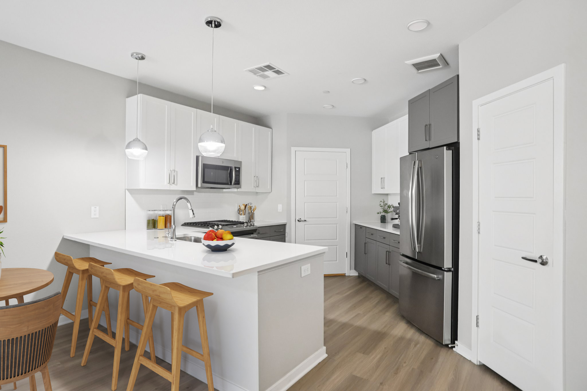Modern kitchen featuring a white island with wooden stools, sleek gray cabinets, stainless steel appliances, and an organized layout. Bright lighting from pendant fixtures and natural light creates a warm atmosphere. A small dining area is visible, adding to the cozy vibe.