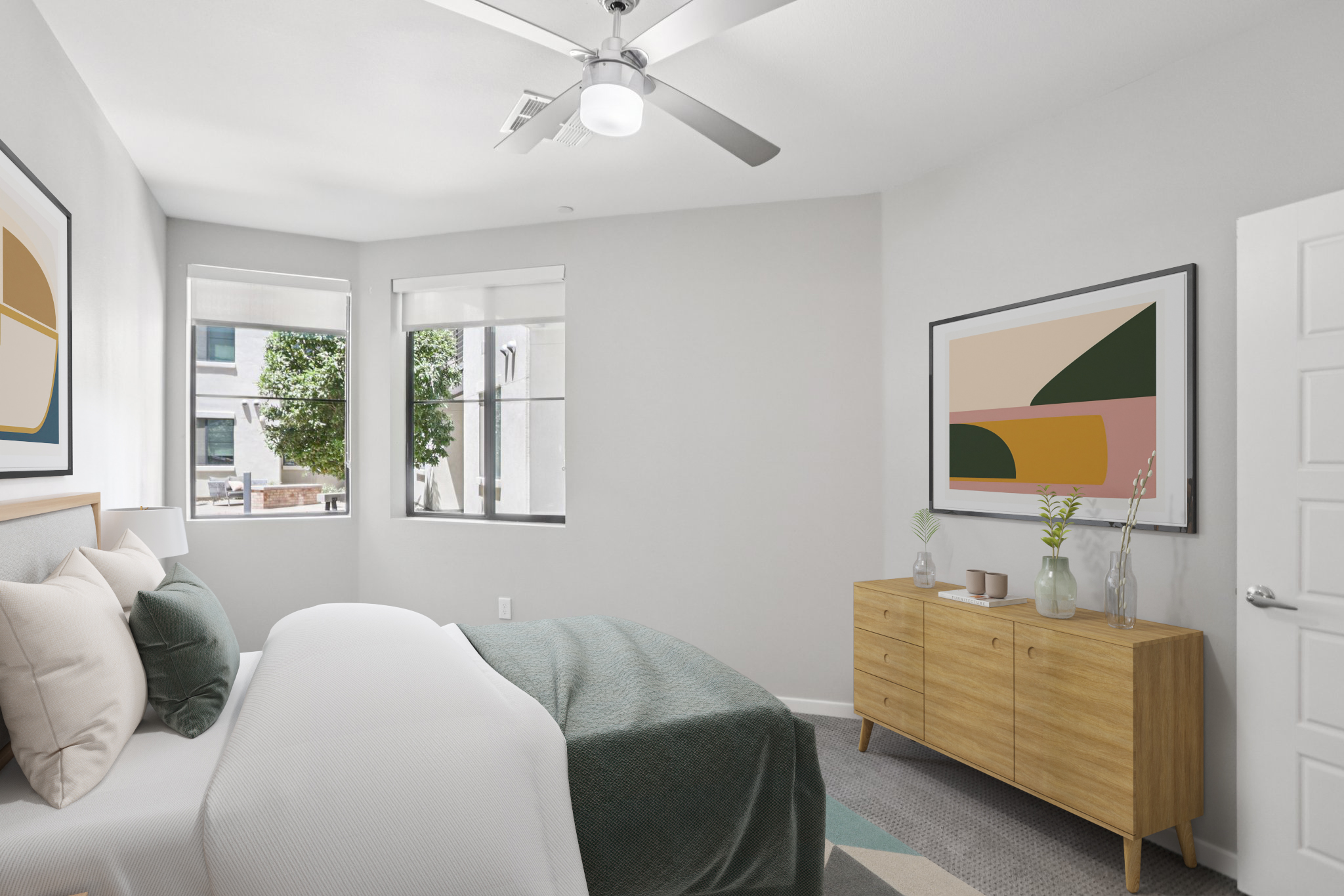 A modern bedroom featuring a bed with a white duvet and decorative pillows, light gray walls, and a wooden dresser. There are large windows allowing natural light, and colorful abstract art hangs on the wall. A ceiling fan is installed above, adding to the room's contemporary style.