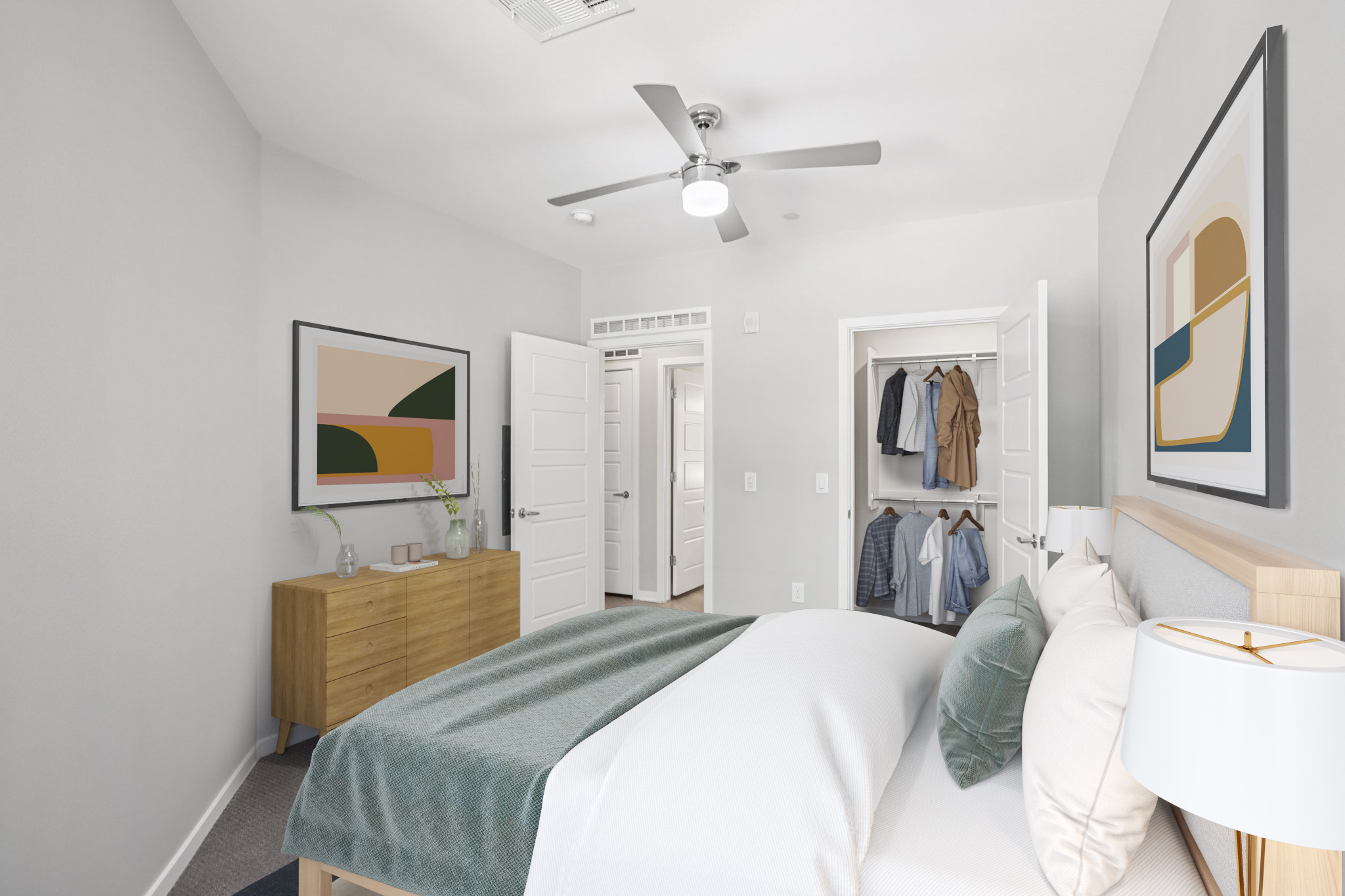 A modern bedroom featuring a bed with a light-colored duvet, a nightstand with a lamp, and a chest of drawers. The walls are painted light gray, and there are two framed abstract artworks. A closet is visible with hanging clothes, and the space is well-lit with a ceiling fan.