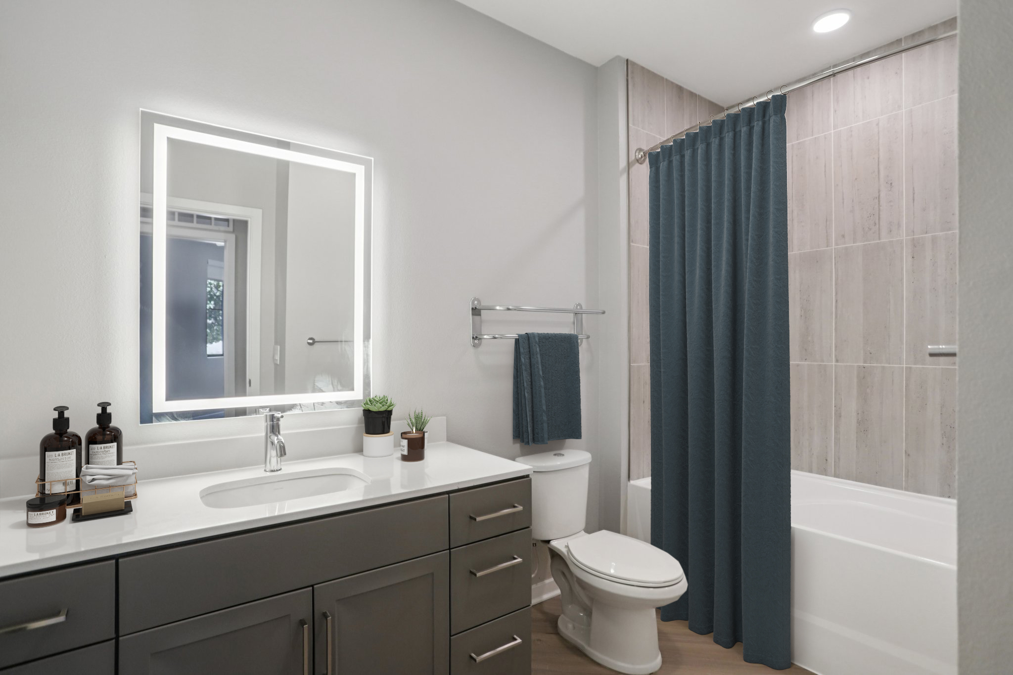 A modern bathroom featuring a gray vanity with a large mirror illuminated by LED lighting, a white sink with a faucet, and neatly arranged toiletries. A blue shower curtain encloses a bathtub, and there are plants and a towel hanging on the wall, all set against light gray walls and wooden flooring.