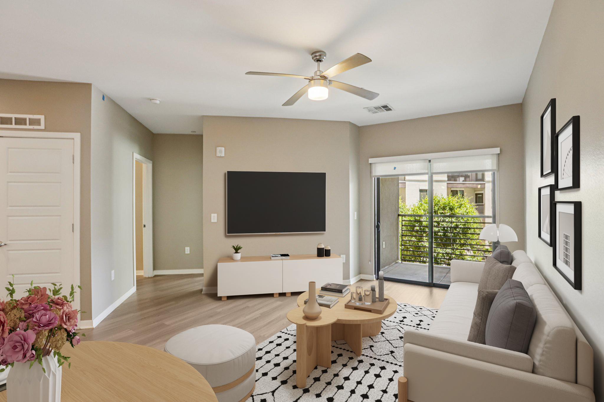 A modern living room featuring a light gray sofa, a round coffee table, and a large flat-screen TV mounted on the wall. There are decorative items on the coffee table and a sliding glass door leading to a balcony with greenery outside. The room is well-lit and has light wood flooring.