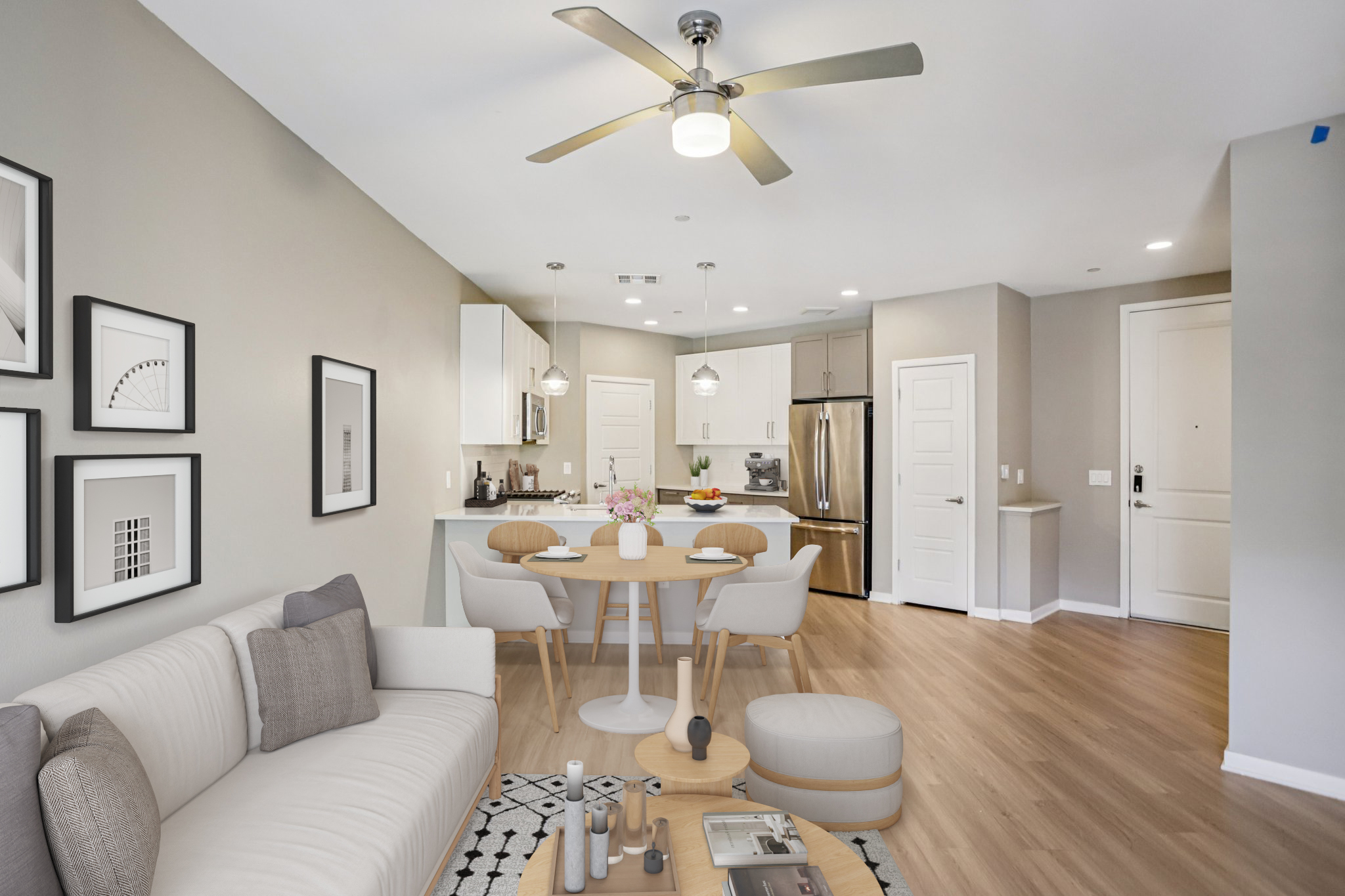 A modern living space featuring a cozy sofa, a round dining table with chairs, and a sleek kitchen with stainless steel appliances. The decor includes framed artwork on the walls and a light color palette, creating a bright and inviting atmosphere. Wood flooring adds warmth to the setting.