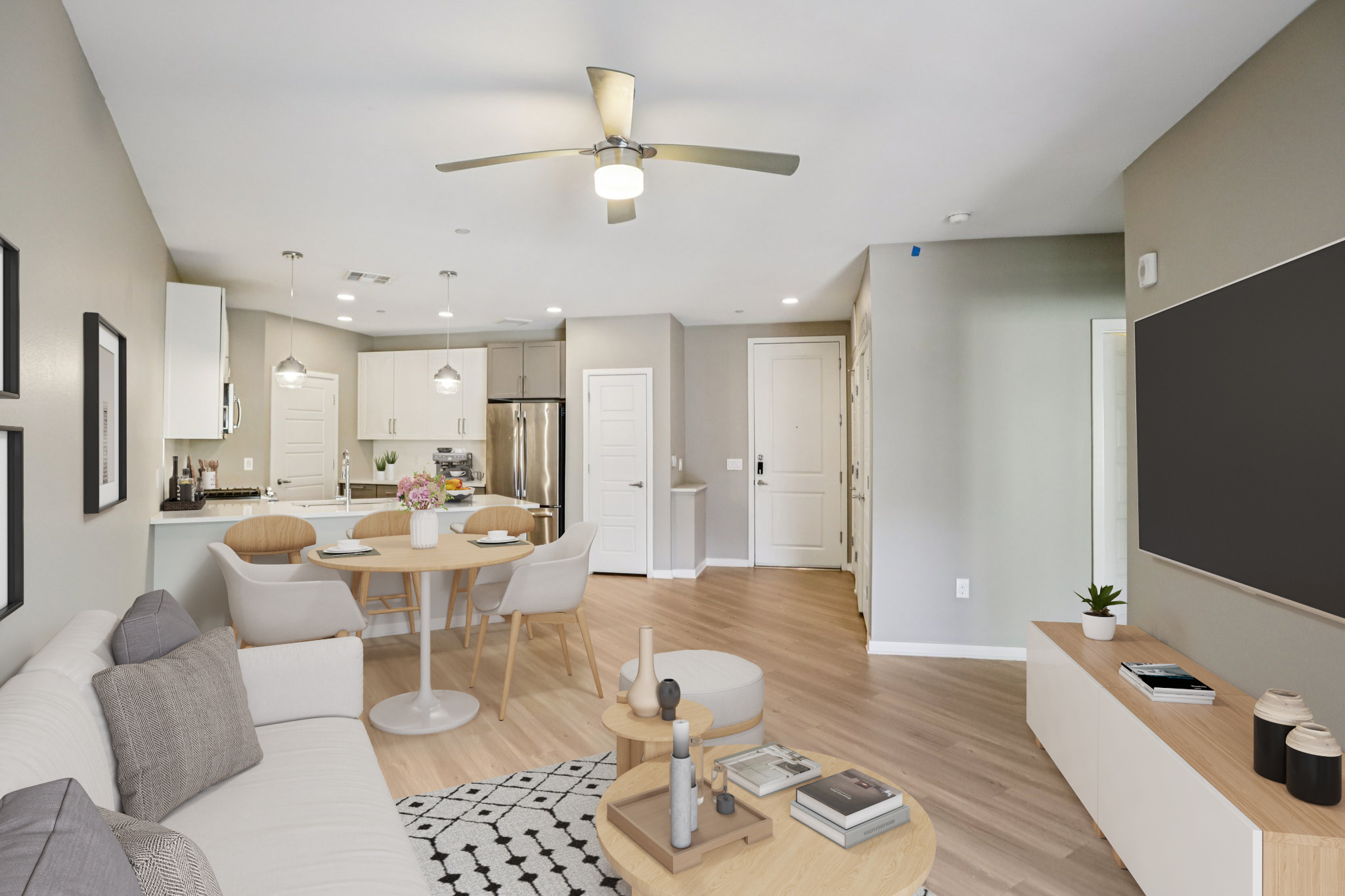 A modern, spacious living area featuring a light-colored sofa, a round dining table with chairs, and a sleek coffee table. The kitchen is visible with white cabinetry and stainless-steel appliances. The floor is light wood, and there are decorative plants and accessories throughout. A ceiling fan adds to the ambiance.