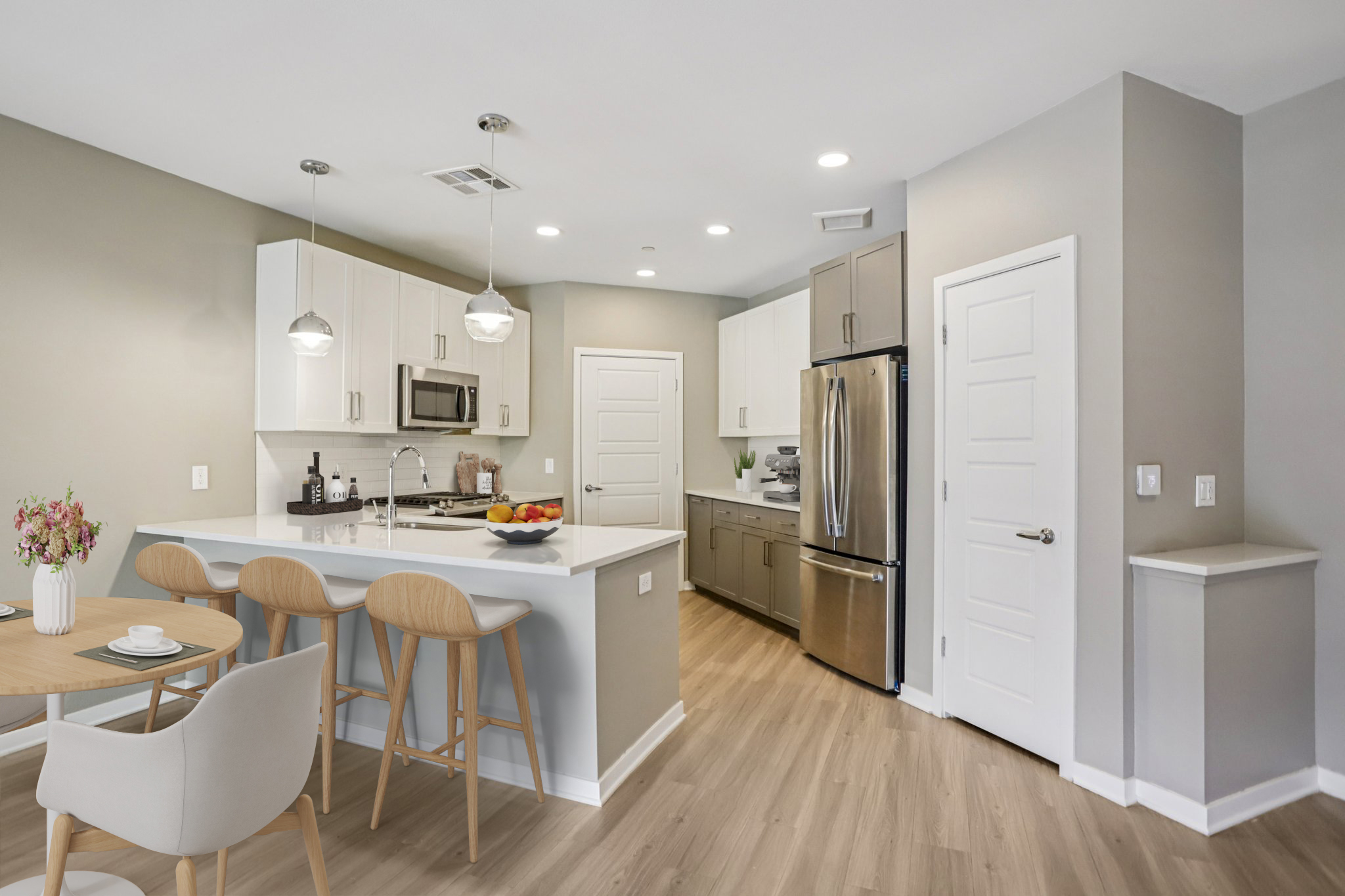 Modern kitchen featuring white cabinetry, stainless steel appliances, and a spacious island with seating. A round dining table with three wooden chairs and a flower vase is situated nearby. The decor includes neutral tones and wooden flooring, creating a bright and inviting atmosphere.