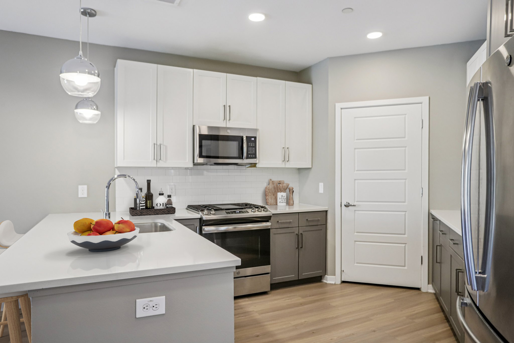 A modern kitchen featuring white cabinets, stainless steel appliances, and a sleek countertop. A bowl of colorful fruits sits in the center, and a door leads to another room. The lighting is bright with stylish pendant lights above the island. The floor has a warm wooden finish, creating a welcoming atmosphere.