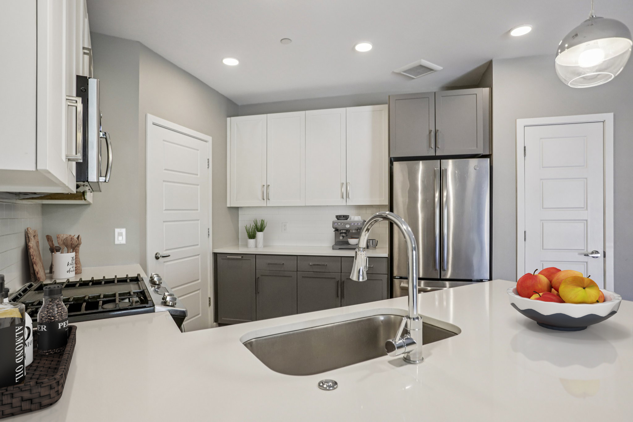 Modern kitchen featuring sleek white and gray cabinetry, a stainless steel refrigerator, a gas stove, and a large sink. A bowl of colorful fruit sits on the countertop, with minimalist decor and a pendant light above. The space is well-lit and organized, displaying a contemporary design.