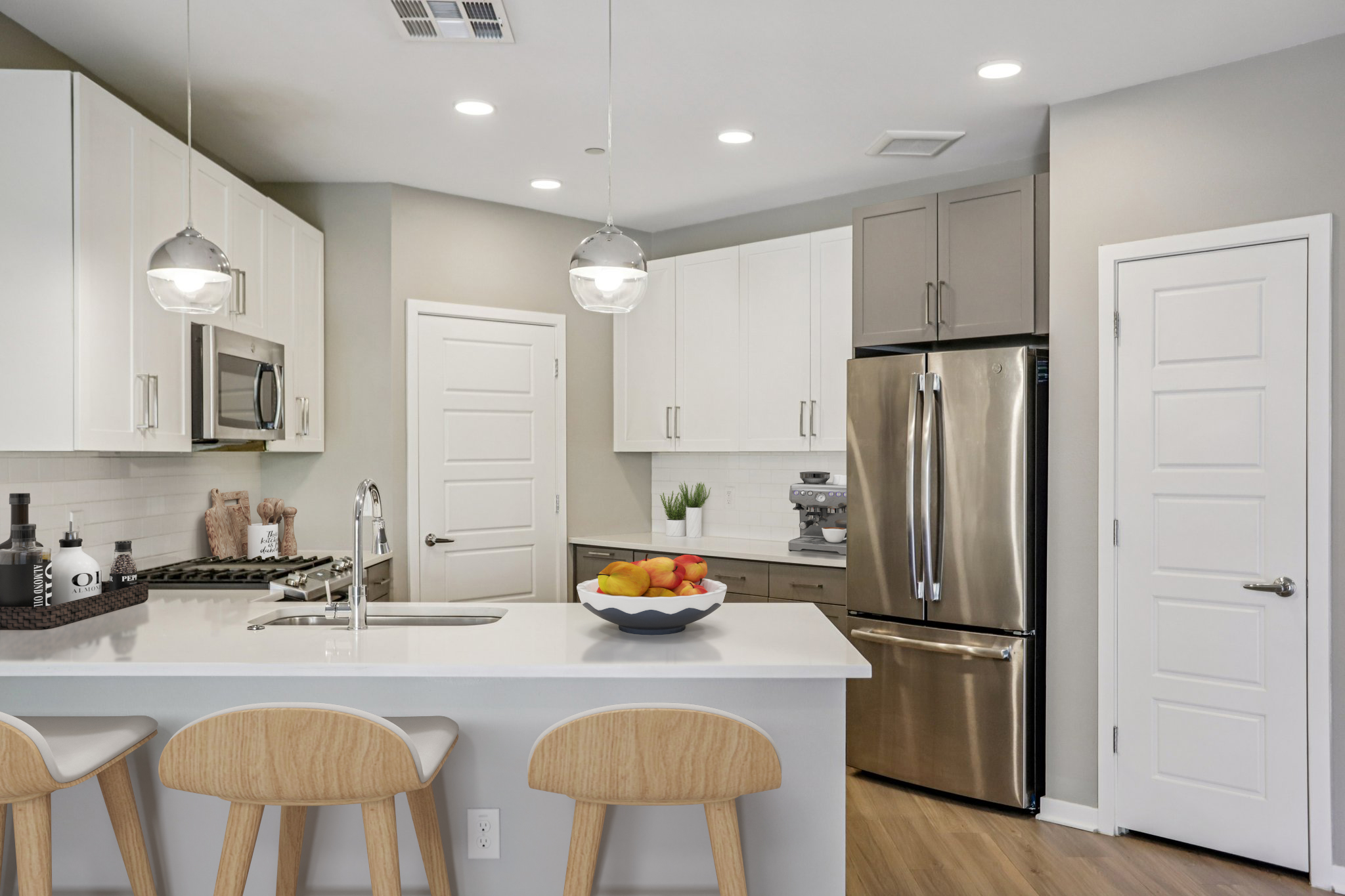 Modern kitchen featuring a sleek design with white and gray cabinets, stainless steel appliances, and a breakfast bar with three wooden stools. A bowl of colorful fruit sits on the counter, and there are recessed lights in the ceiling, contributing to a bright and inviting atmosphere.