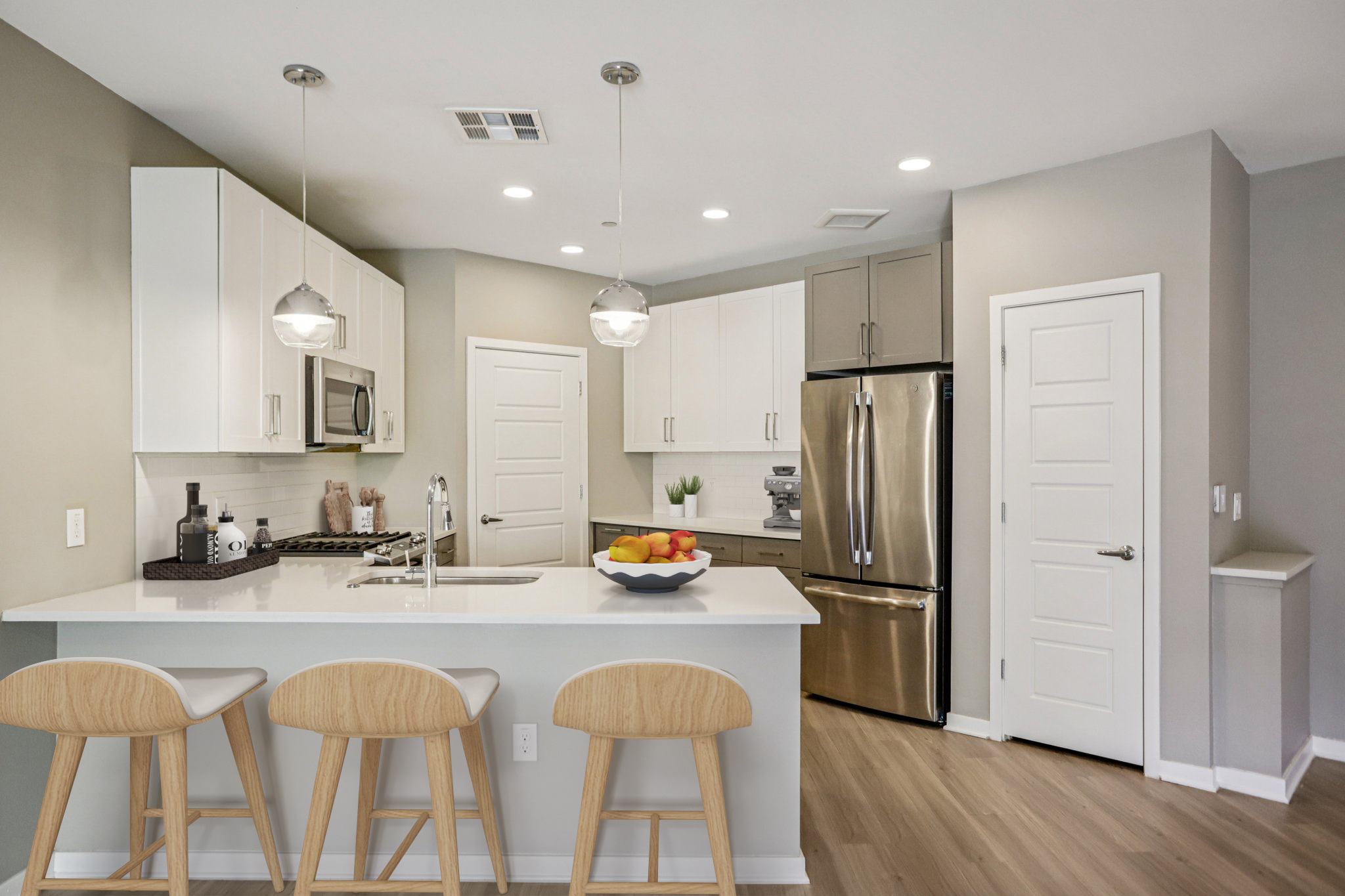 Modern kitchen with white cabinetry, stainless steel appliances, and a large island with three wooden bar stools. The island features a decorative bowl of colorful fruit. The walls are painted in a light gray tone, and there is natural light coming in from a nearby window.