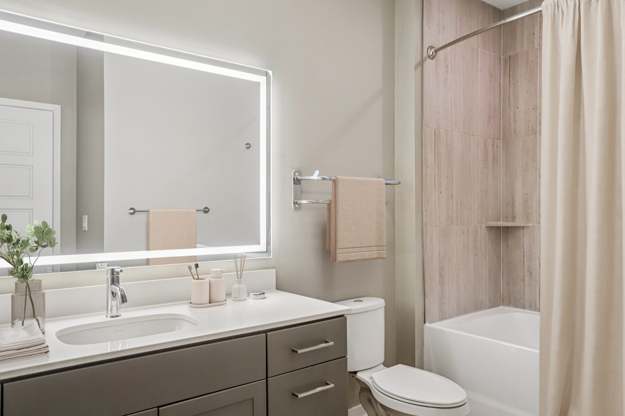A modern bathroom featuring a white countertop with a sink, a large illuminated mirror, and a shower/tub combo. Soft beige towels hang on a rack, and a small plant adds a touch of greenery. The walls are a light color, enhancing the airy atmosphere.