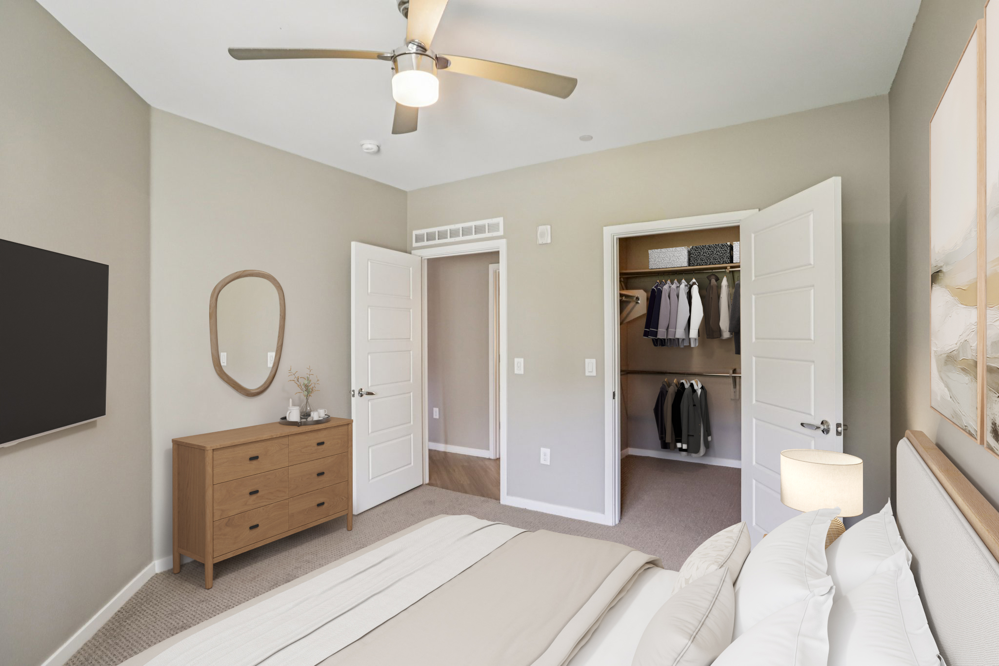 A well-decorated bedroom featuring a bed with white linens, a wooden dresser, and a wall-mounted TV. There are two open doors leading to a closet and another room, with a mirror hanging on the wall. A ceiling fan provides ventilation, and soft lighting creates a cozy atmosphere.