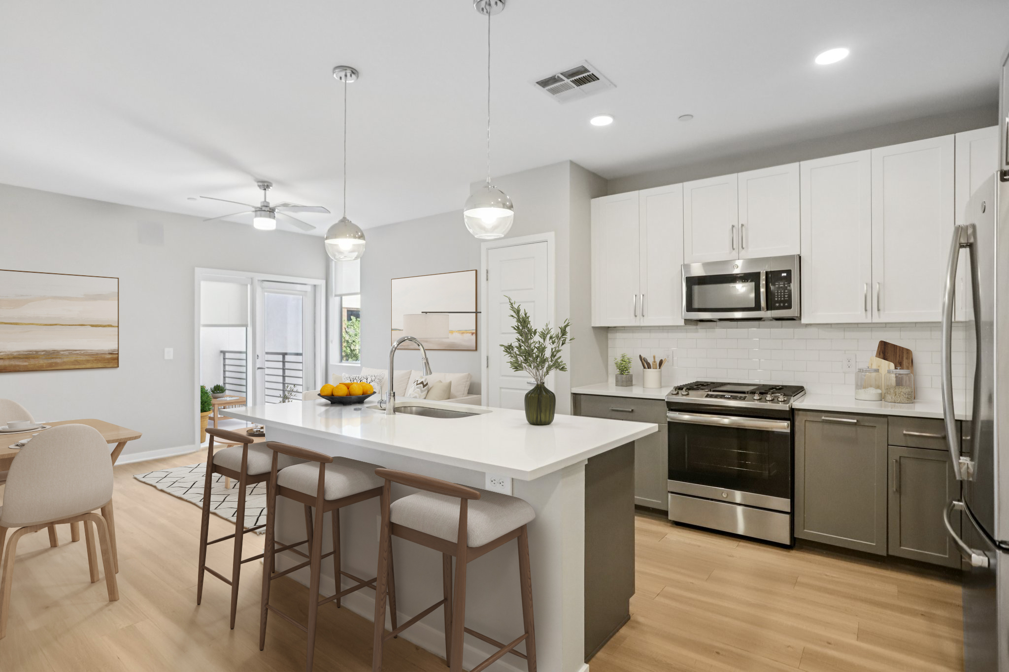 Modern kitchen featuring a white island with bar seating, stainless steel appliances, and light wood cabinetry. A dining area with round chairs and artwork is visible in the background, along with natural light coming from large windows. Minimalist decor adds to the contemporary aesthetic.