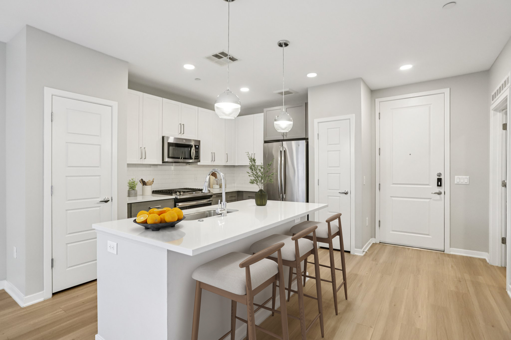 Modern kitchen featuring white cabinets, stainless steel appliances, and a large island with bar stools. Bright and airy with pendant lighting, the space includes a bowl of oranges on the counter and a door leading to the hallway. Neutral color palette adds a clean and contemporary feel.