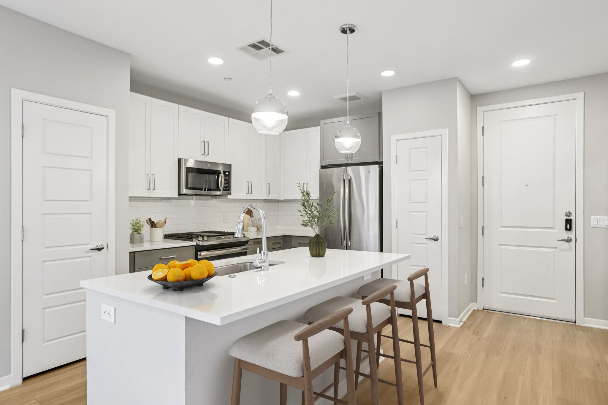 A modern kitchen featuring sleek white cabinetry, stainless steel appliances, and a large island with three bar stools. A bowl of oranges sits on the island, and there's a door leading to an entrance. The space is well-lit with pendant lights and has a clean, contemporary design.