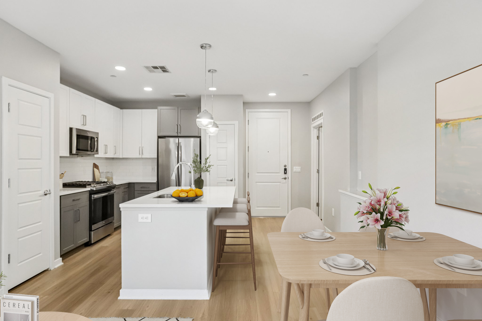 A modern kitchen and dining area featuring white cabinetry, stainless steel appliances, and a central island with bar stools. A wooden dining table is set with plates and a floral centerpiece, while a bowl of oranges rests on the counter. The space is brightly lit with neutral-colored walls and flooring.