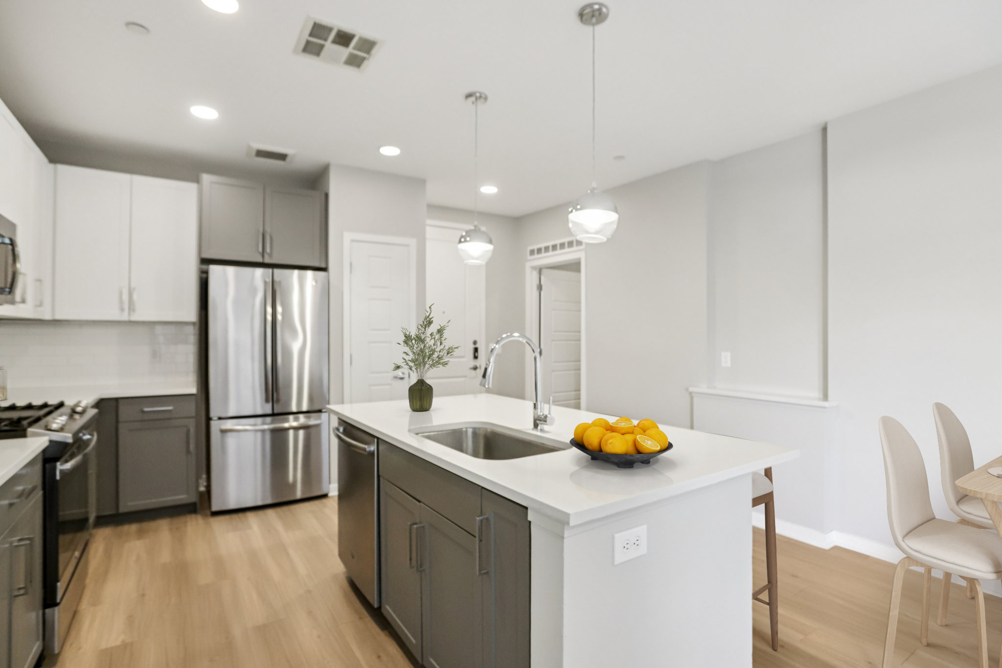 A modern kitchen featuring a central island with a sink, a bowl of oranges, stainless steel appliances, and white cabinets. The space has light wood flooring and bright, neutral-colored walls, with pendant lights hanging above the island, creating a spacious and inviting atmosphere.