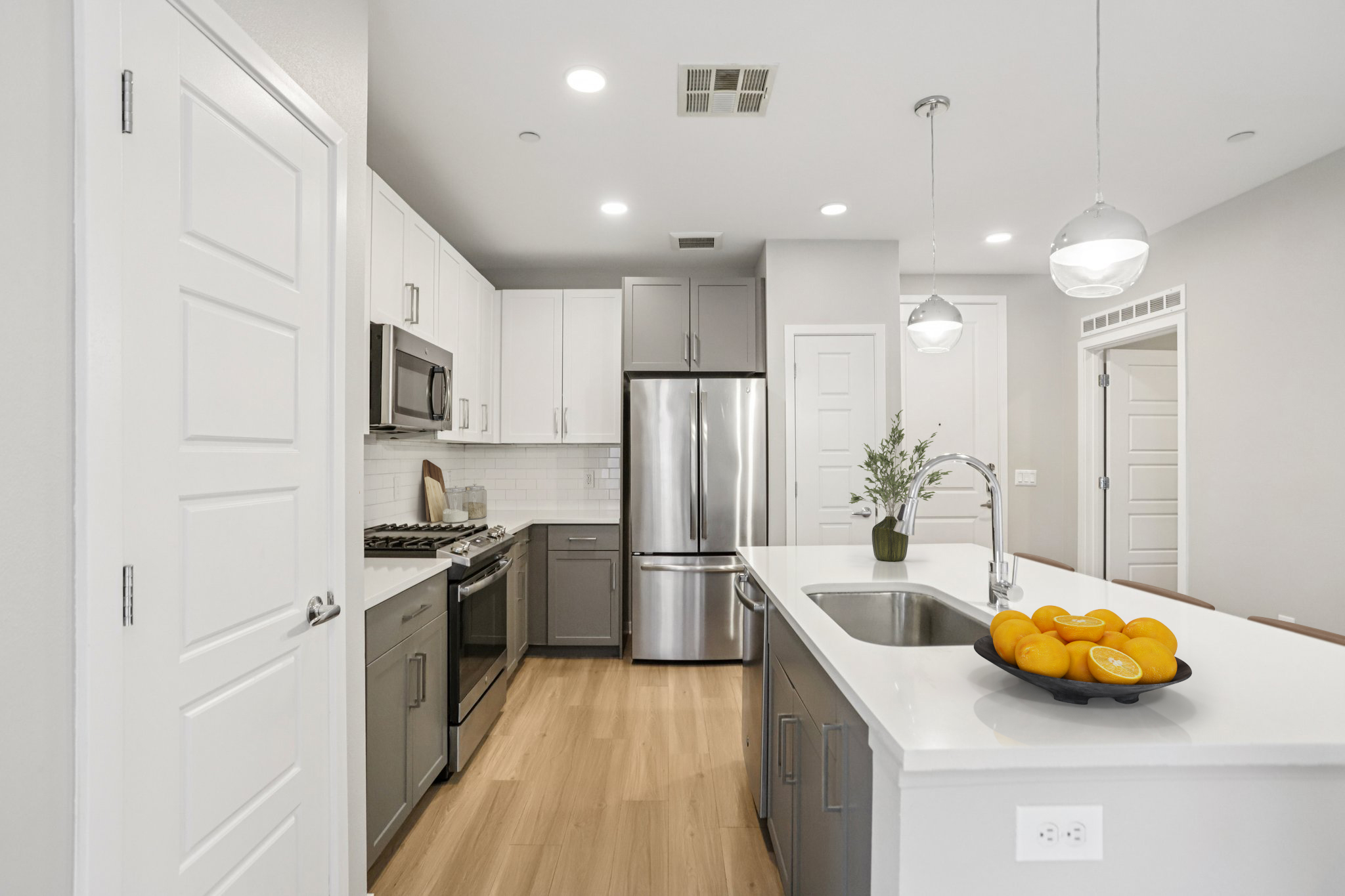 Modern kitchen featuring stainless steel appliances, a spacious island with a bowl of oranges, light wood flooring, and a minimalist design. Cabinets in gray and white, complemented by a tile backsplash and ample natural light. The area is open and inviting, with a doorway visible in the background.
