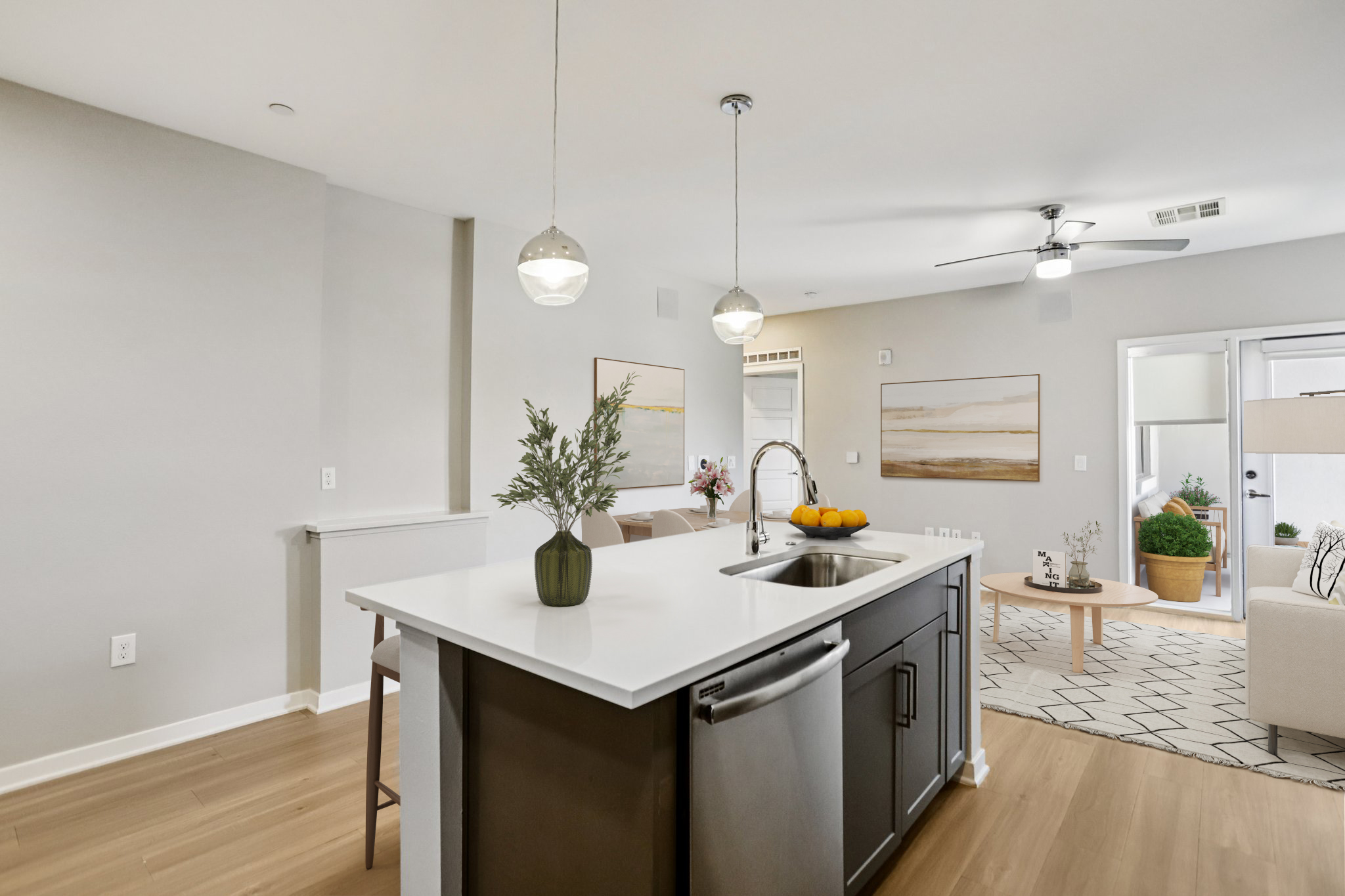 A modern kitchen featuring a central island with a sink, surrounded by stylish decor. On the island, there's a vase with greenery and a bowl of oranges. Light wood flooring complements the neutral walls and contemporary artwork in the background. The space is bright and inviting, with a ceiling fan and open layout leading to a living area.