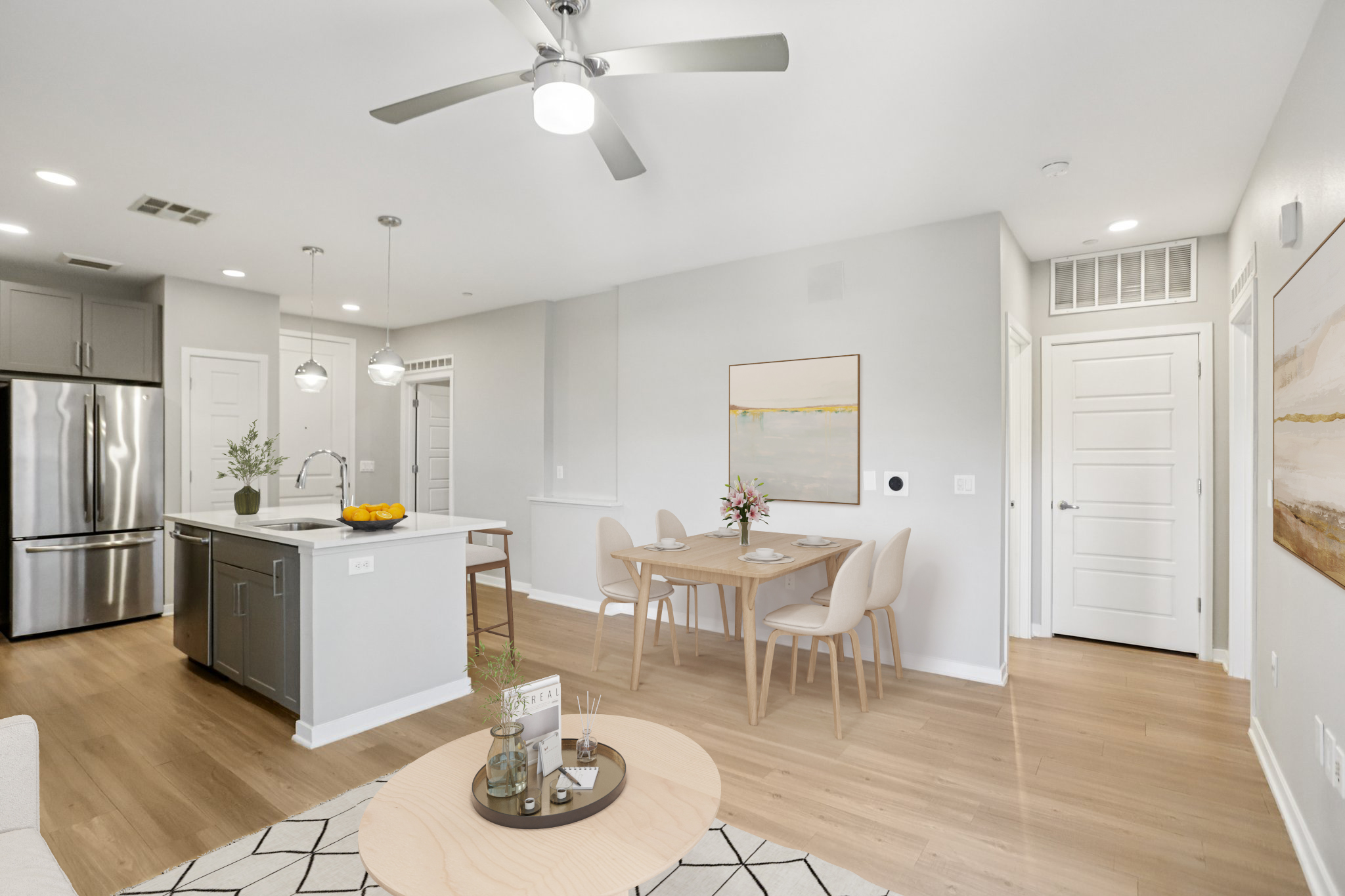 A modern open-concept kitchen and dining area featuring stainless steel appliances, a large island with a fruit bowl, a dining table with light wood chairs, and minimalistic decor. The light gray walls and warm wooden floors create a bright, inviting atmosphere.