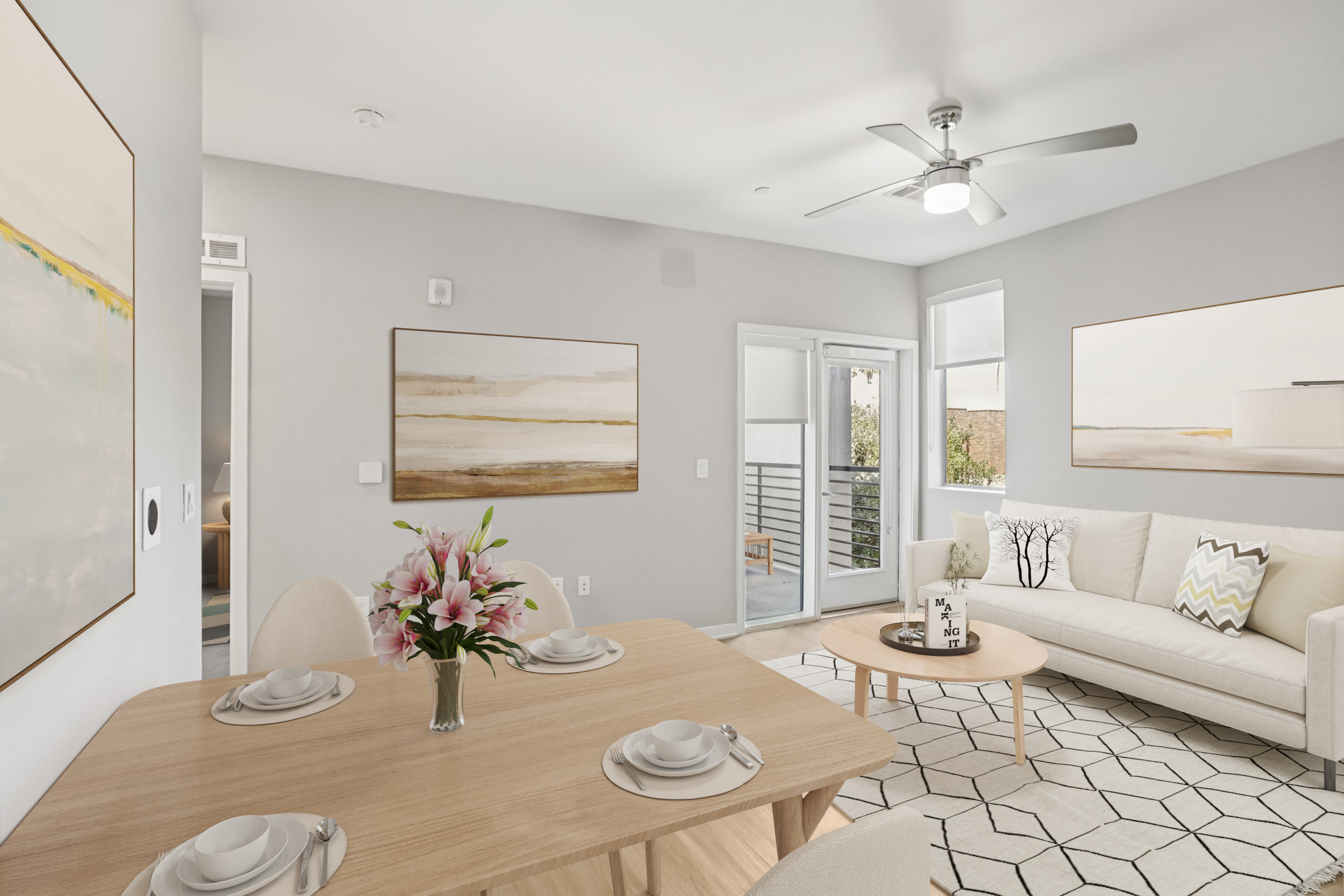 A modern living space featuring a light-colored sofa with decorative pillows, a wooden dining table set for four, and a vase of pink flowers. Two large abstract artworks decorate the walls, and large windows provide natural light. A sliding glass door opens to a balcony.