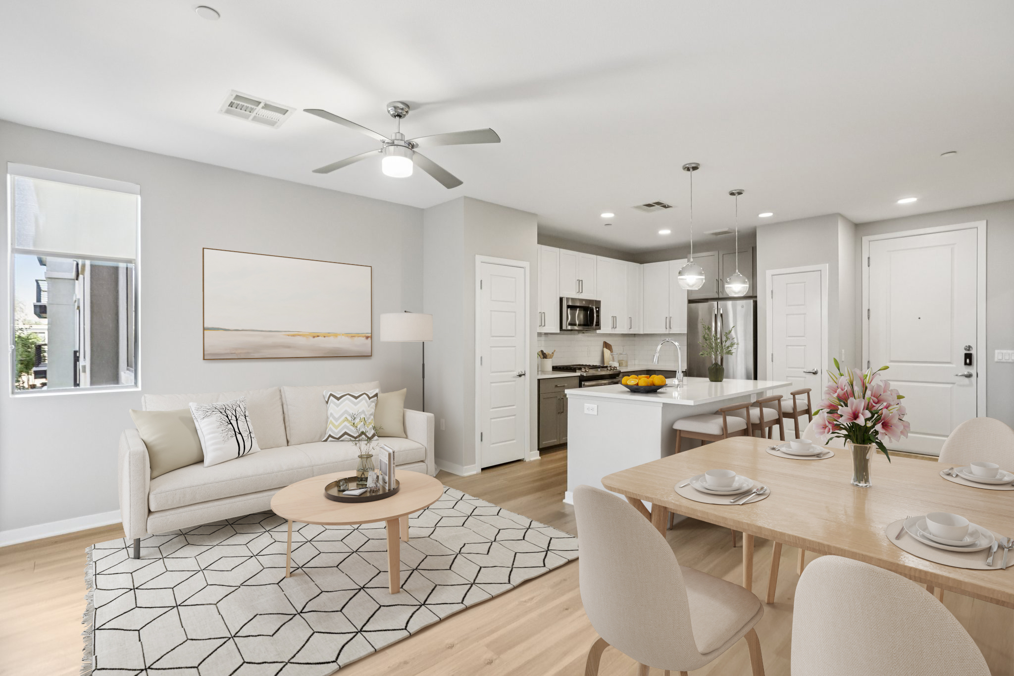 Bright, modern living area featuring a light gray wall, a comfortable beige sofa with decorative pillows, and a round coffee table. The open-concept kitchen is visible, showcasing white cabinetry and stainless steel appliances. A dining table is set for four, with a vase of flowers and a patterned rug on the wooden floor.