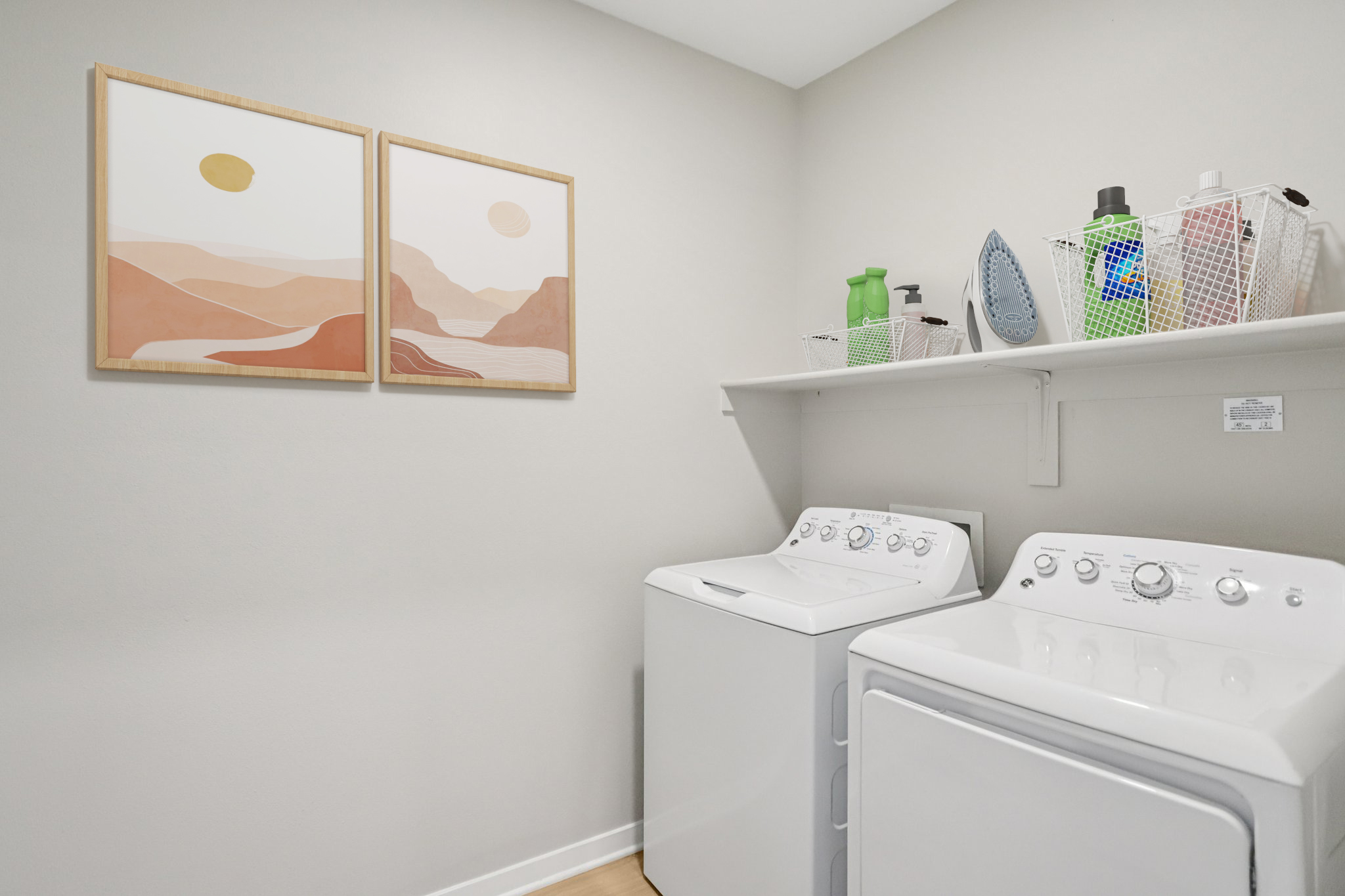 A modern laundry room featuring a white washing machine and dryer side by side. Above them, a shelf holds cleaning supplies and an iron. The walls are light gray, adorned with two framed abstract landscape paintings in warm tones, adding a stylish touch to the space.
