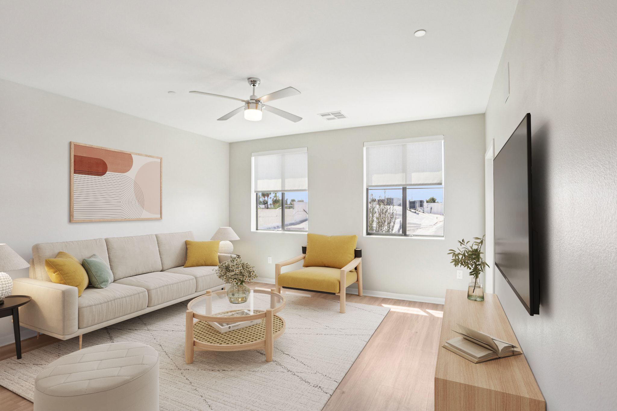A bright and modern living room featuring a comfortable beige sofa with yellow accent pillows, a round coffee table with a vase of flowers, and a cozy yellow armchair. There are two windows with shades that allow natural light in, a ceiling fan, and a minimalist TV stand against the wall, creating a welcoming atmosphere.